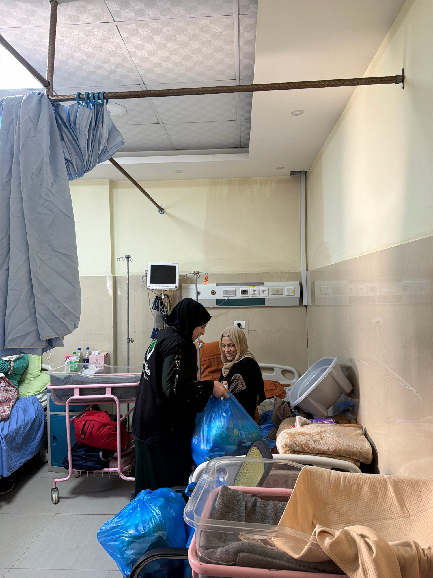 The image depicts an indoor scene, likely a hospital room, in Gaza City, Palestinian Territories. Two women are present. One woman, wearing a black hijab and a dark abaya with green accents, stands to the left. She is handing a large blue plastic bag filled with items to the second woman. The second woman, wearing a light-colored hijab and a dark outfit, is seated and smiling. She is reaching out to receive the bag.
The room appears to be cluttered with various objects. To the left, a pink metal baby crib with wheels is visible, containing some bedding. A red bag and other items are placed underneath it. Medical equipment, including a monitor and an IV stand, are mounted on the wall. Several plastic bags, some blue and some clear, are scattered around the floor and on a transparent container. Blankets and other textiles are piled up in the foreground and on a nearby surface. The walls are painted a light color, and the ceiling is tiled. A metal rod, likely part of a curtain rail system, extends across the upper portion of the image. The overall impression is one of a functional yet somewhat disheveled environment.