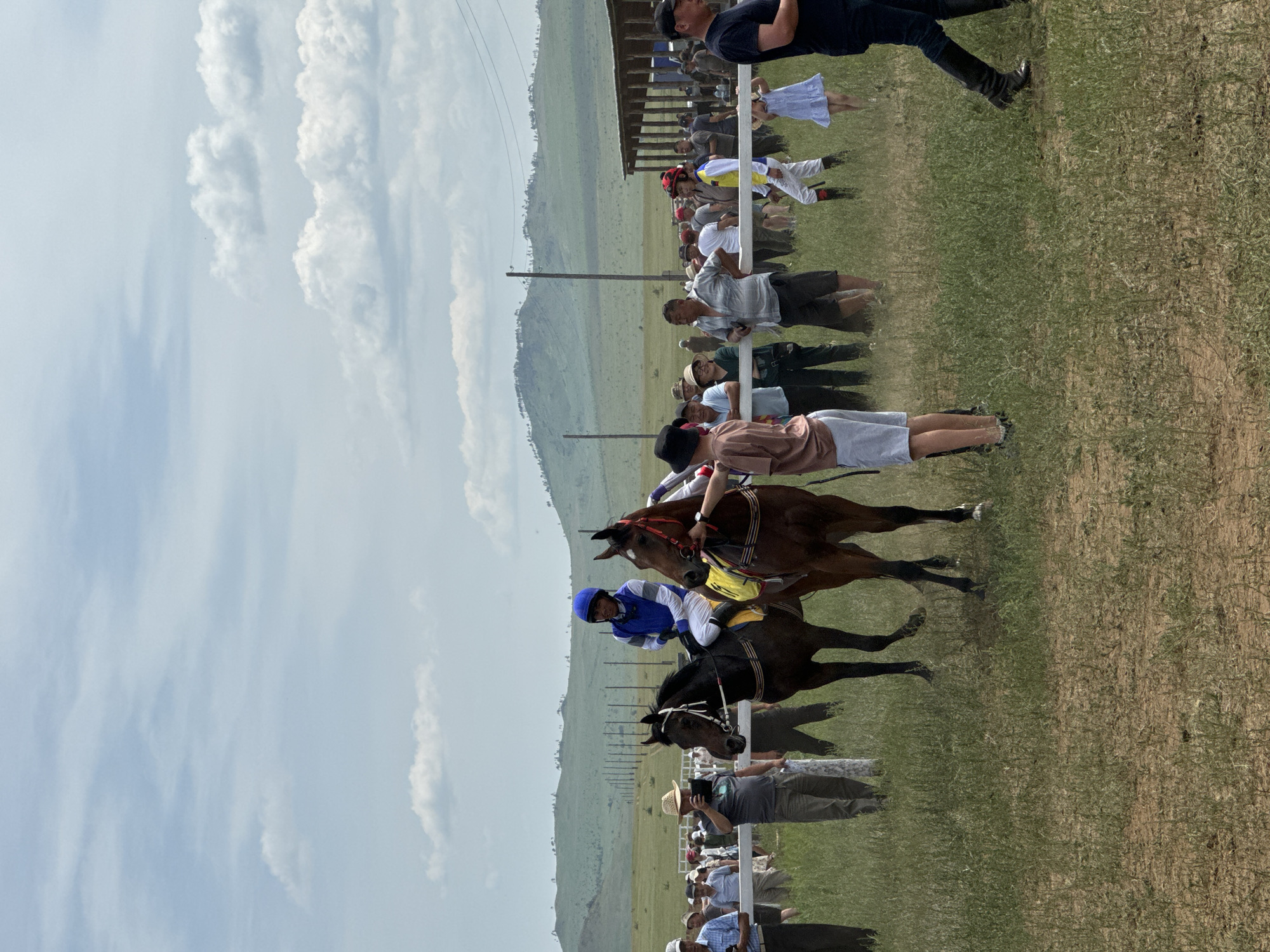 The image depicts a horse race event in Orongoi, Russia. The scene features several people gathered on a grassy field, with two horses and their jockeys in the foreground. One jockey, wearing a blue and white uniform, is mounted on a dark brown horse with a yellow saddle pad. Another jockey on a similar horse is partially visible to the right. Spectators, dressed in casual attire, are standing behind a white fence. In the background, a wooden structure, likely a stable or grandstand, is visible, along with rolling hills and a line of utility poles. The sky is partly cloudy.