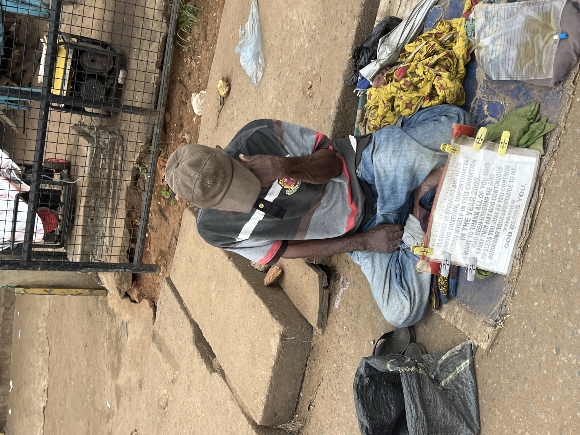 The image shows a man sitting on a concrete surface, likely outdoors in Nkpor, Nigeria. He is wearing a baseball cap, a striped t-shirt, and jeans. To his right, a white sign with text written in black is clipped onto a piece of fabric. The text on the sign reads: "A WAH MAK OF 75 YEARS OLD, TWAD ELOQUENT IN SPEAKING BUT HARD WORKING SOMEONE, UNFORTUNATELY FOUND MYSELF IN THIS CONDITION IN THE YEAR 2000. I WANT TO THANK ALL OF YOU WHO HAD BEEN HELPING ME AND I PRAY THAT MAY GOD CONTINUOUS TO BLESS YOU JUST AS YOU CONTINUE RENDERING YOUR ASSISTANCE TO ME. THANKS GOD BLESS YOU."
Next to the sign are some yellow clips and a green cloth. Behind him, there are piles of fabric, including a yellow cloth with red star patterns, a plastic bag, and what appears to be a medical bag. To the left of the man, a metal fence encloses a generator. The ground is made of concrete and dirt, with some debris scattered around. The lighting suggests it is daytime. The man's posture and the context of the sign imply he is likely a beggar seeking assistance.