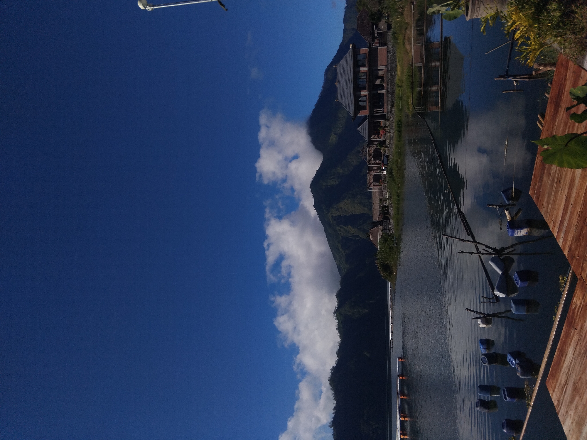Here is a description of the image:
The image depicts a landscape featuring a body of water, likely a lake, flanked by a mountain range and buildings. The sky is blue with scattered white clouds, some of which are near the mountain peaks. The water reflects the sky and the buildings. Along the shoreline, there are structures and what appears to be a wooden dock or platform. There are small, blue and white items floating in the water, possibly buoys. A street light pole is visible in the top left corner. The scene appears to be set in a rural or semi-rural location, blending natural and man-made elements.