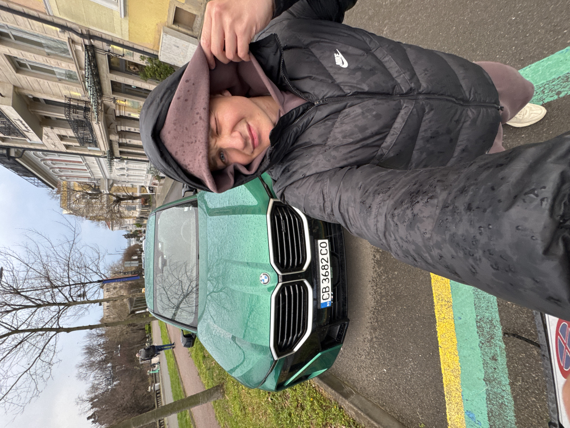 The image shows a person taking a selfie with a green BMW car in Burgas, Bulgaria. The person is wearing a black Nike puffer jacket with a hoodie pulled over their head, and a light purple sweatshirt underneath. The car has a Bulgarian license plate, CB 3682 CO. The background features buildings lining the street and bare trees along a sidewalk. The street has yellow and green painted lines. The weather appears to be wet, with rain droplets visible on the car and the person's clothing.