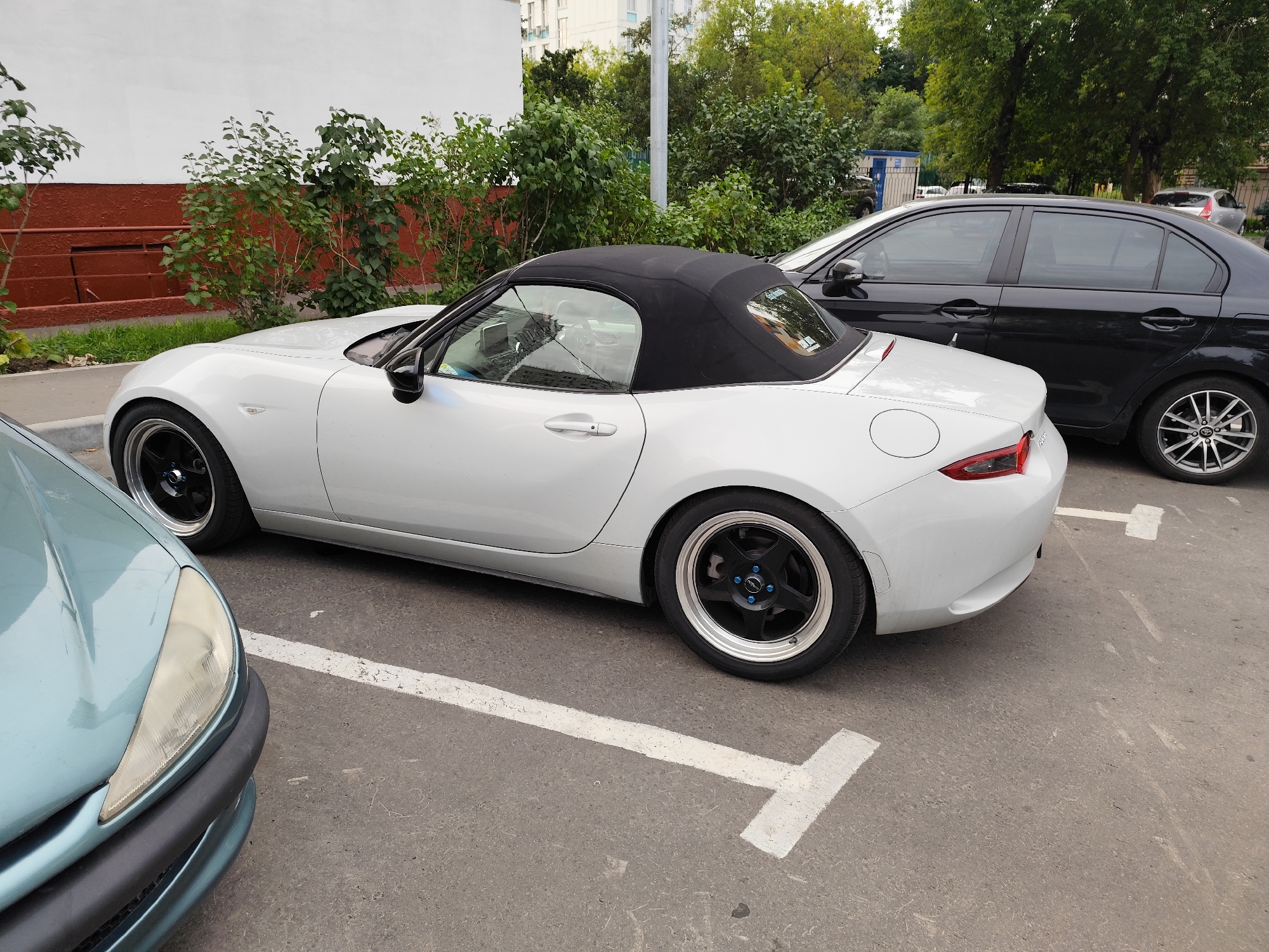 A white convertible Mazda Miata and a black sedan are parked on a paved surface marked with white parking lines. The convertible is in the foreground, with its black soft top up. The black sedan is parked to the right and behind the convertible. A light blue vehicle's front bumper and headlight are visible on the far left of the image. In the background, there is a white building, green trees, and a light pole. The scene appears to be a parking lot in a residential area, possibly in Moscow, Russia.
