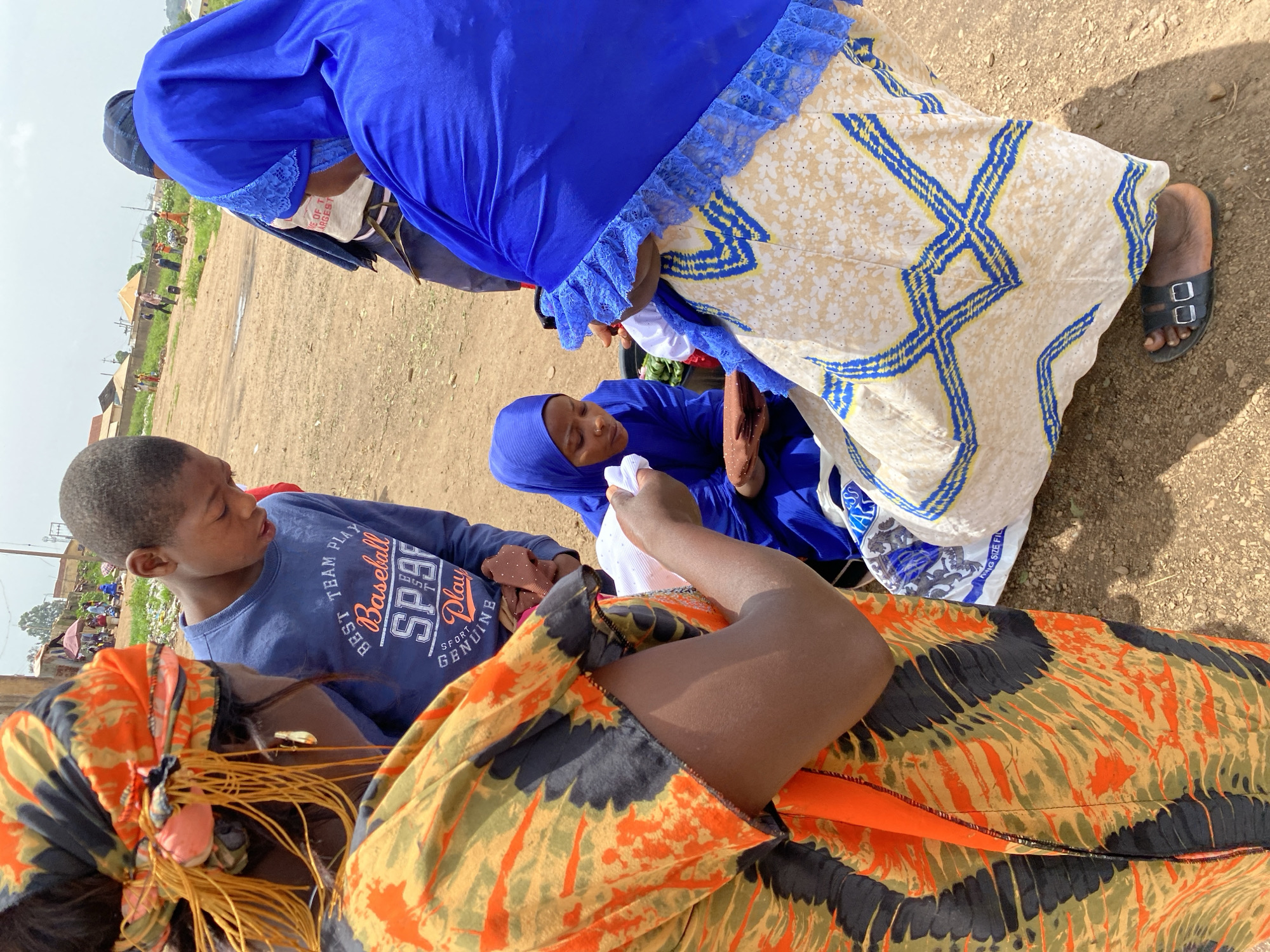 This media file shows a group of people, including children and adults, outdoors in what appears to be a market or community gathering. The scene is set on a dirt ground with some vegetation and distant buildings visible. The people are dressed in traditional clothing, with vibrant colors and patterns. Some are wearing headscarves or hijabs.
The focus is on a young boy wearing a blue sweatshirt with the text "BEST TEAM PLAY Baseball SP9 Play GENUINE" printed on it. He is looking to the side, seemingly engaged in a conversation or observing something. Nearby, a woman is holding a baby, both covered by a blue garment. Another person, possibly an adult, is wearing a flowing cream and blue patterned dress.
The overall impression is one of community and everyday life. The lighting suggests it is daytime, and the weather appears to be warm and possibly sunny, given the shadows. The presence of market stalls and multiple people in the background hints at a lively environment. The text on the boy's shirt is clearly legible and adds a detail to his attire. The people's expressions are not distinctly visible due to the angle and resolution, but the overall mood seems relaxed.