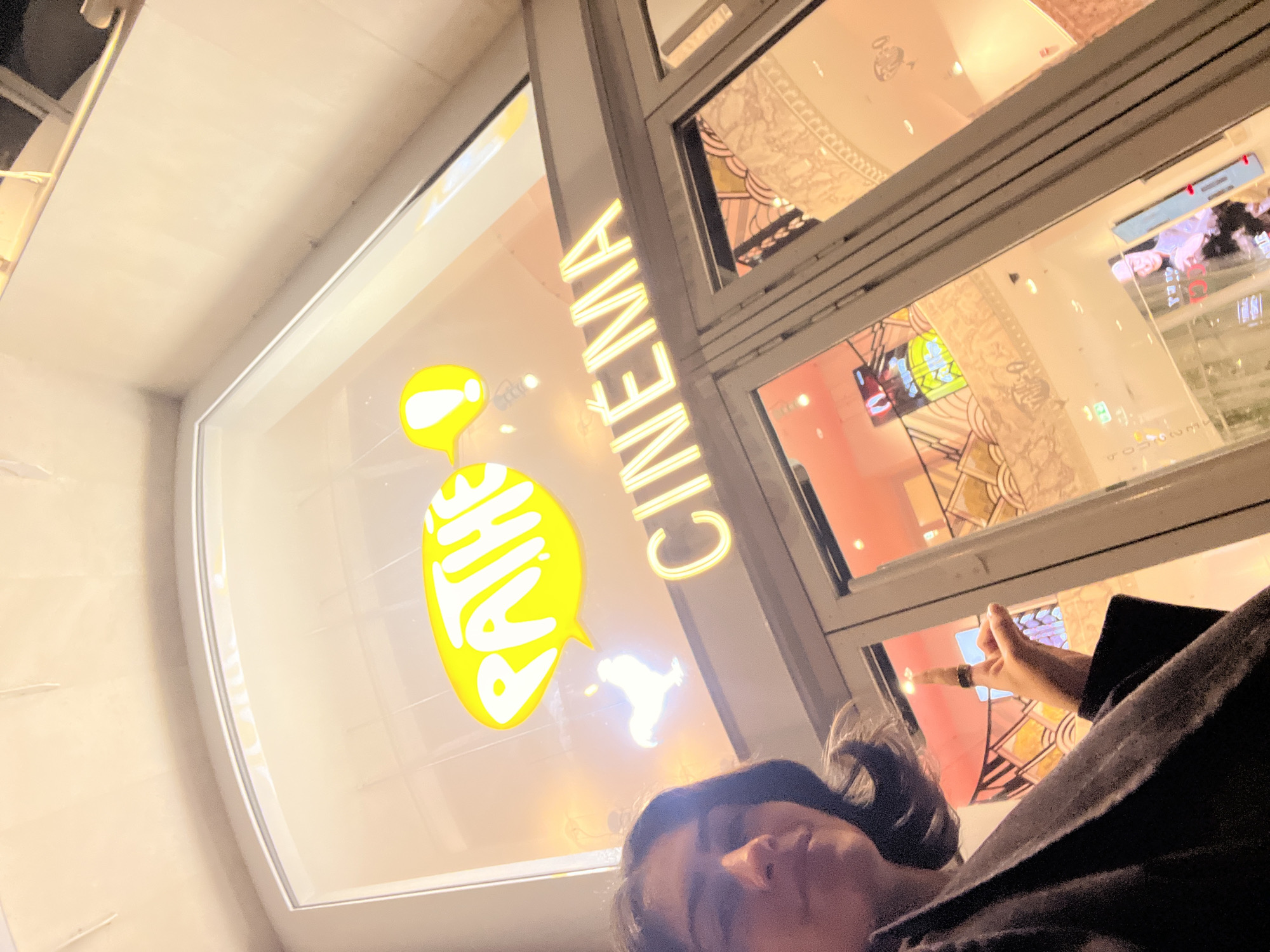 A low-angle outdoor shot shows a woman lying down, possibly on the ground, in front of a building with "PATHE CINEMA" signage. The woman is positioned in the lower center and is partially obscured by the perspective, with her face visible. Her clothing is dark. The "PATHE" logo is in the form of stylized yellow speech bubbles. The "CINEMA" lettering is vertical and illuminated in yellow. Reflections on the glass windows show elements of the building's interior, including decorative patterns and interior lighting. The location is likely in Paris, France, due to the presence of "Pathe Cinema," a prominent cinema chain operating in France.