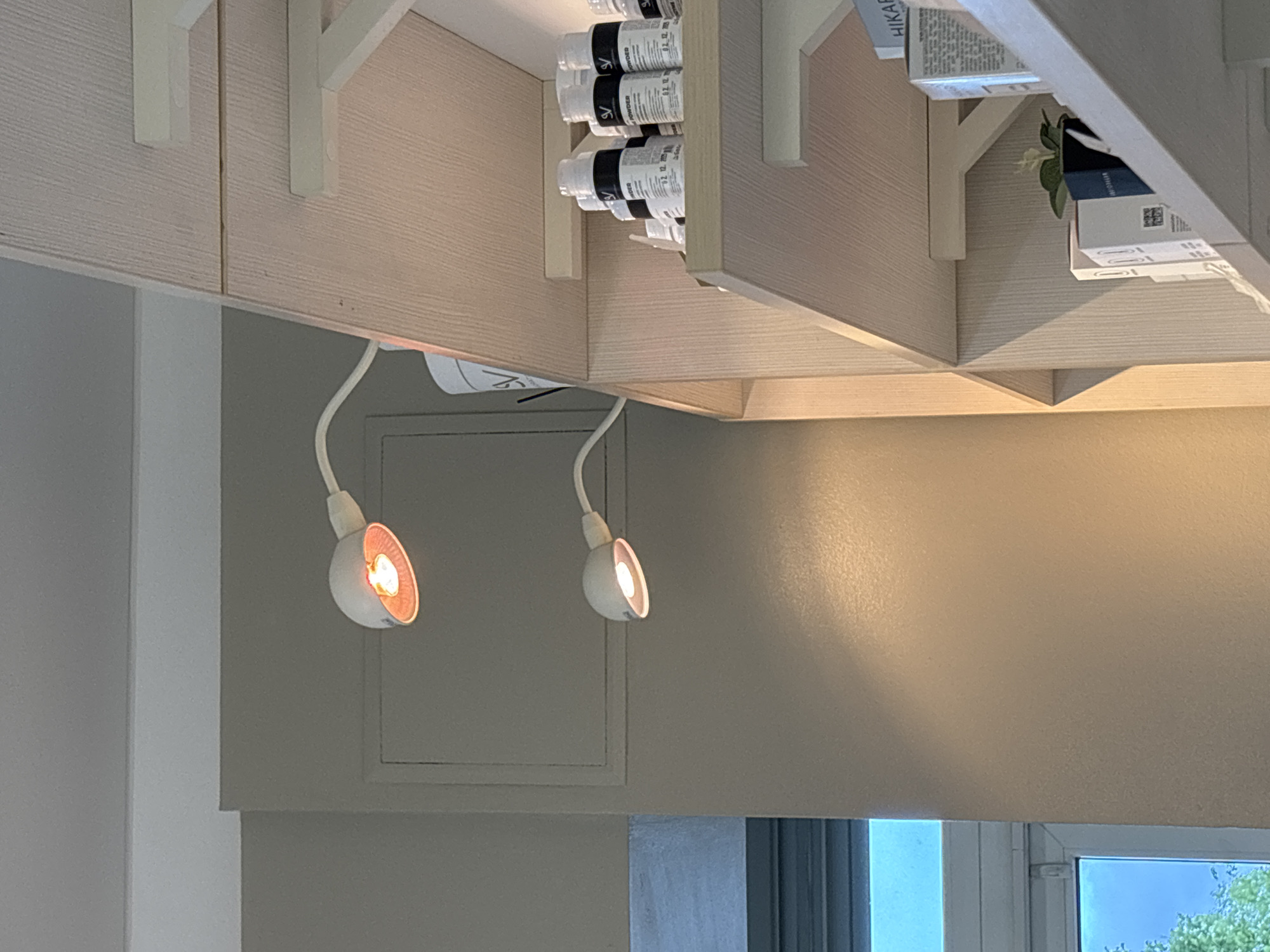This image depicts an upside-down view of a room interior, likely a kitchen area. Visible elements include overhead cabinets in a light wood-grain finish, supported by brackets. One cabinet contains containers labeled "SV POWDER" and another has a box labeled "HIKARI." Below the cabinets, two small spotlights with flexible necks are illuminated, casting a warm glow. A gray wall with a framed access panel is visible behind one of the lights. A window with a partial view of the outside, including a tree, is also present. The exact location within the city of Unknown, Russia, cannot be determined from the given image.