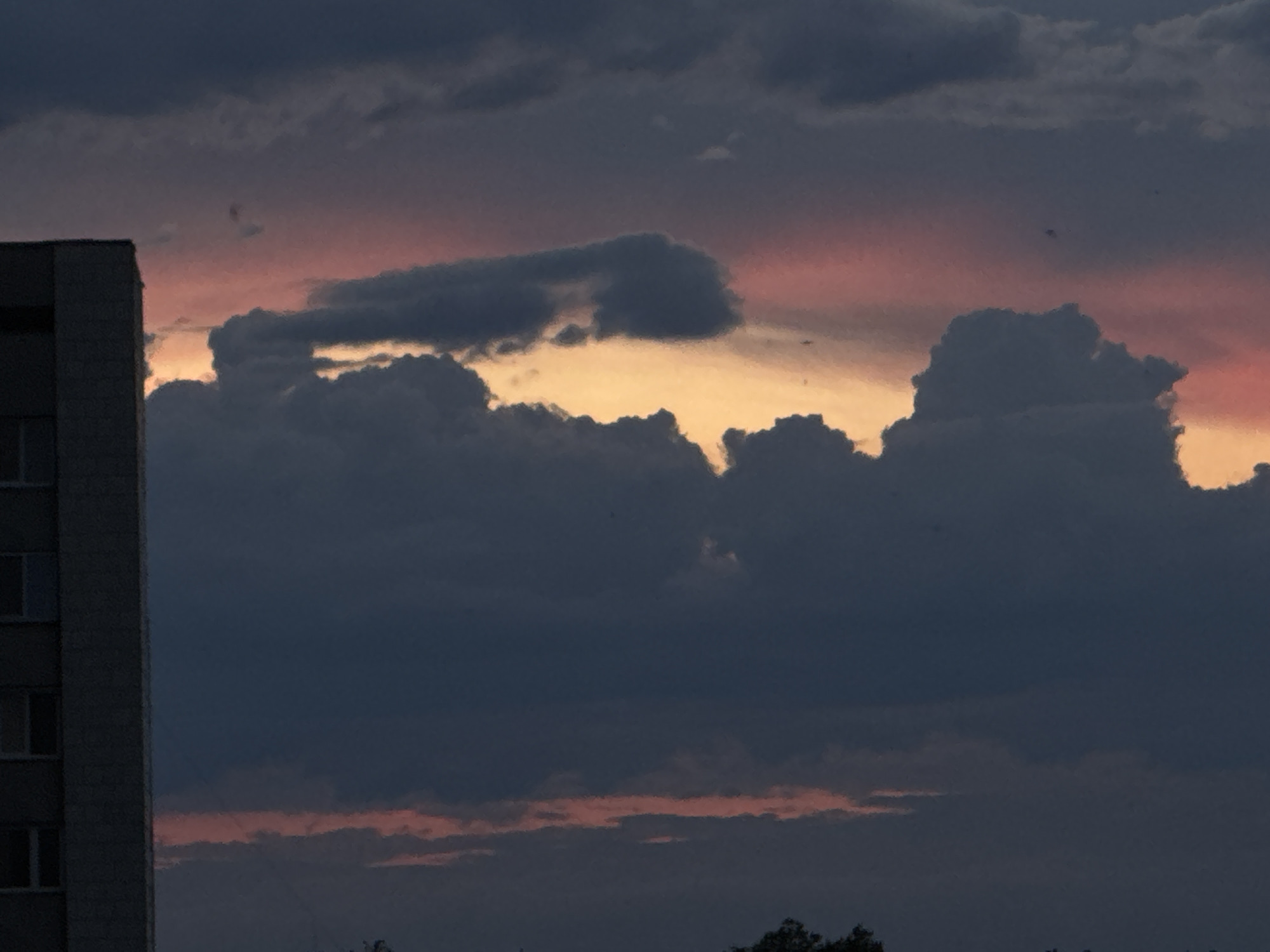 The image captures a dramatic sunset over Tiraspol, Moldova. The sky is filled with dense, dark clouds tinged with hues of orange, pink, and yellow where the sun's rays break through. A portion of a tall, utilitarian building, likely an apartment block, is visible on the left side of the frame, its windows dark and indistinct. The building's facade appears to be made of concrete. The lower part of the image shows the silhouettes of trees against the dimly lit sky. The overall atmosphere is somber and somewhat ominous, characteristic of a stormy sunset.