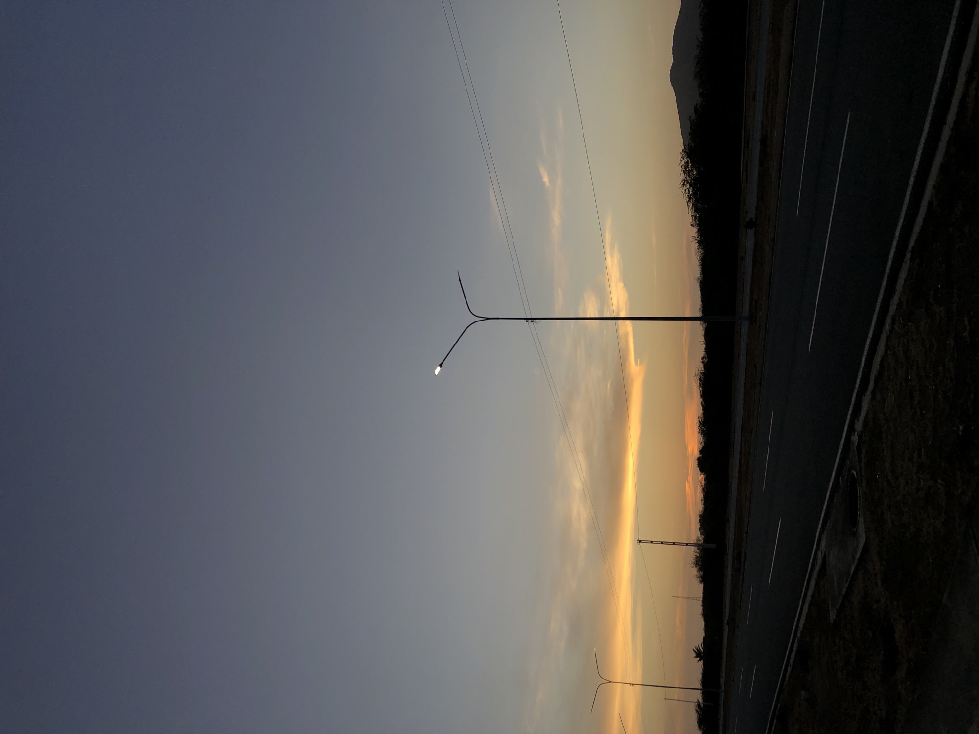 The image shows an evening landscape with a road running along the right side. To the right of the road is a dark, grassy area, and beyond that, a line of trees. In the distance, there is a hill or mountain range. The sky transitions from a deep blue on the left to a golden, sunset-like color towards the right, with wisps of clouds visible. Utility poles and lines run across the sky, and several streetlights are visible along the road. The scene appears to be in Altagracia, Venezuela.