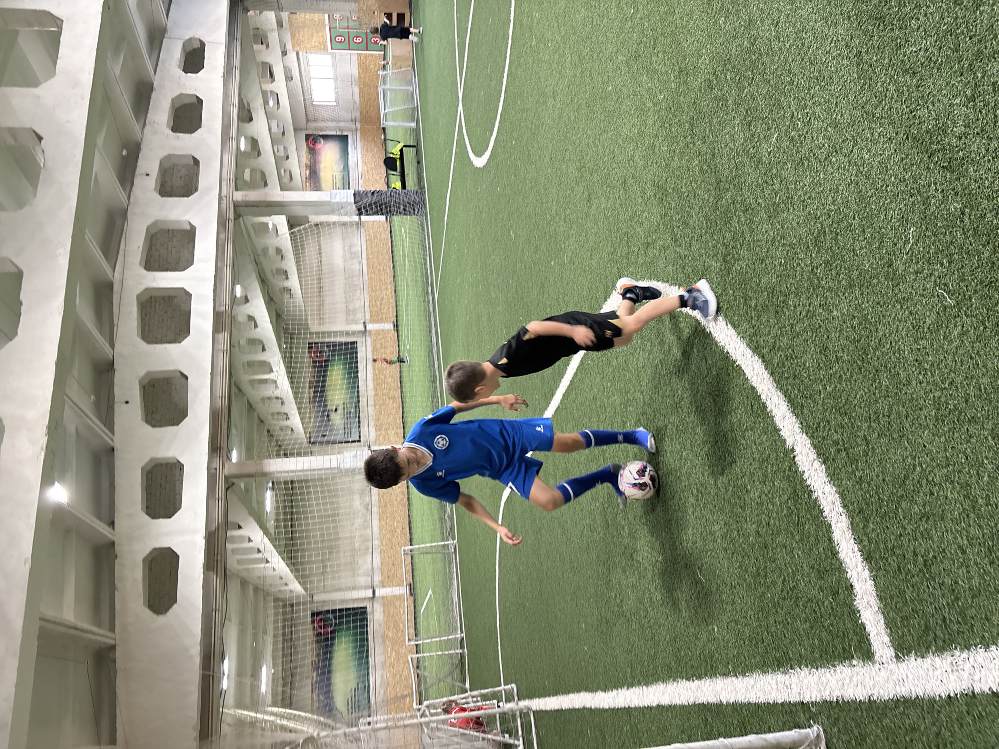 The image shows two young boys playing soccer on an indoor turf field. The boy in the foreground is wearing a blue soccer jersey and shorts, with blue socks and cleats. He is positioned to the left of a soccer ball, with his right foot about to kick it. The second boy, in the background, is wearing a black t-shirt and shorts, and also has cleats. He is in motion, appearing to be running.
The setting is an indoor sports facility with a green turf field marked with white lines. The field is enclosed by netting and has goals at either end, though only one appears to be fully visible. The background shows the interior of the building, with concrete pillars, elevated walkways, and what appear to be posters or murals on the walls. There is a scoreboard visible in the upper part of the image with the numbers 3 and 6 displayed. The lighting suggests it is daytime or a well-lit indoor space.
The overall mood is one of active play and likely friendly competition, common for children's sports activities. The image is taken from a slightly elevated, angled perspective, giving a dynamic view of the boys and the field. The location context provided is Chaikovsky, Russia, which suggests this is a local sports facility. There is no indication of weather conditions as it is an indoor scene.