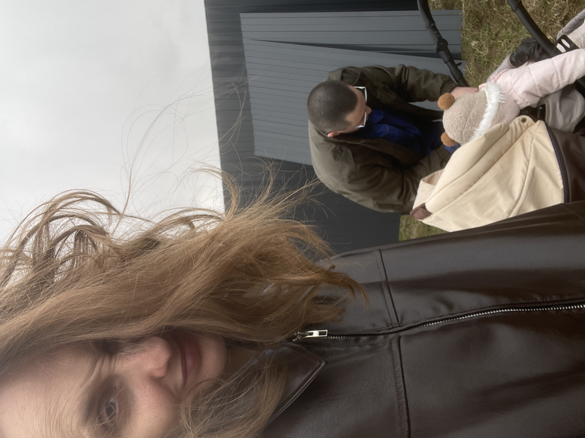 The image, taken in Podshivalovo, Russia, is a selfie capturing a family outdoors under an overcast sky. In the foreground, positioned to the left, is a young woman with long, flowing light brown hair that appears wind-swept. Her face is partially visible, showing one eye and the side of her mouth, with some facial lines suggesting a smile or active expression. She is wearing a dark brown or black leather-look jacket, the zipper of which is visible on the right.
In the mid-ground, a man with short hair and glasses is seen leaning over a baby stroller. He is wearing a dark green jacket over a blue shirt. His attention is directed downwards towards the stroller. Inside the stroller, a baby is visible, dressed in a light pink jacket and wearing a light-colored hat with two prominent ear-like features, possibly a teddy bear design. The stroller itself is light beige and dark brown.
The background features a modern-looking building with dark horizontal siding, suggesting an industrial or contemporary architectural style. To the right of the man and stroller, a patch of green grass is visible, indicating an outdoor, possibly park-like or suburban setting. The overall atmosphere is casual, and the wind-blown hair of the woman suggests breezy conditions. There is no visible text in the image.