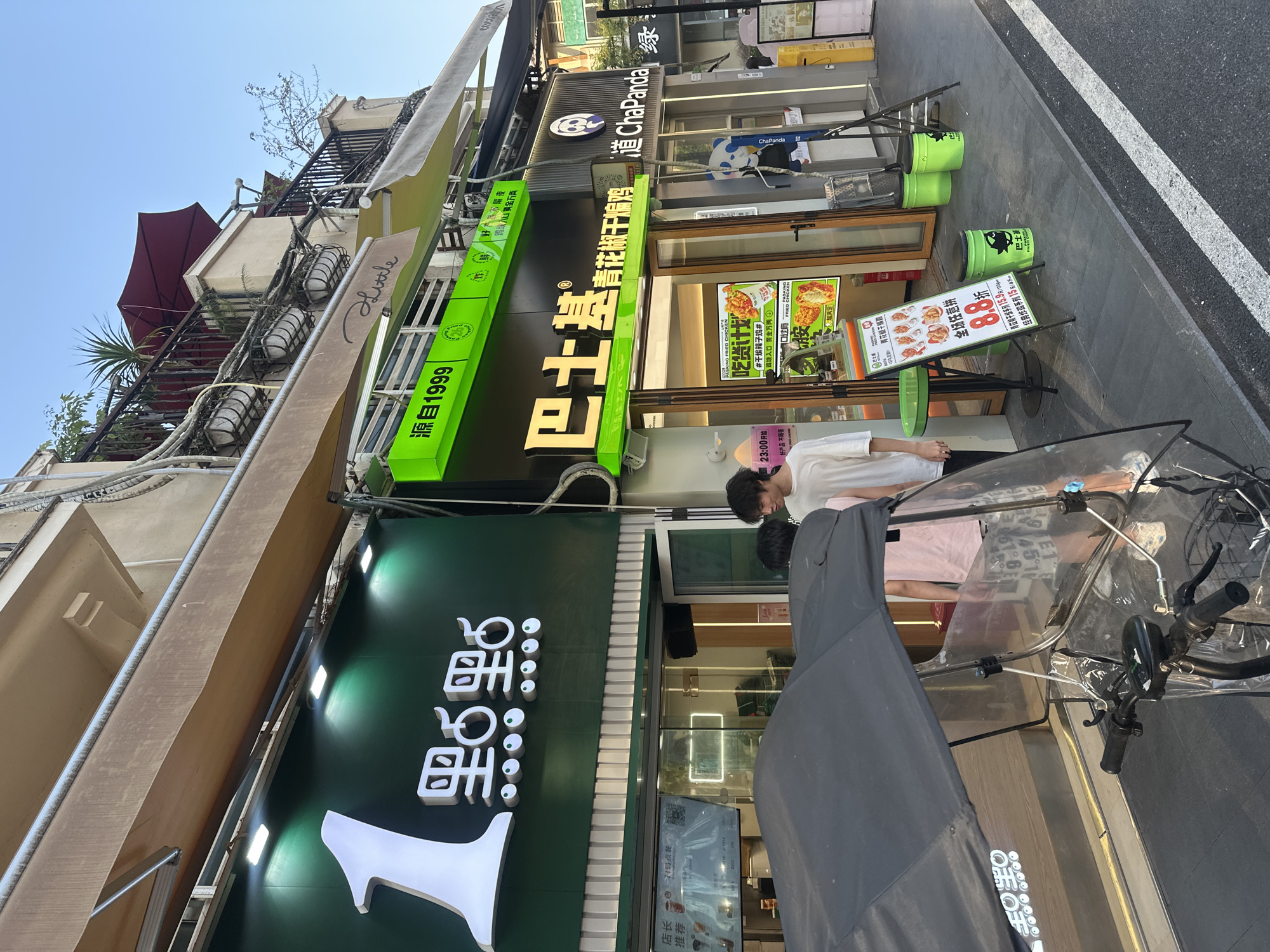 This image captures a vibrant daytime street scene in Chongqing High-tech Zone, China, under a clear blue sky. The setting is a commercial area with multiple shops lining a sidewalk.
The main subjects are the storefronts, two individuals, and a bicycle. From left to right, a shop with a green awning and a cursive "Little" sign, along with "源自1999" (Founded in 1999) is partially visible. The most prominent shop is "青花椒干煸鸡" (Green Peppercorn Dry-Fried Chicken), featuring large golden Chinese characters on a black and green signboard. Its window displays promotions like "PASKING FRIED CHICKEN," "吃货计划" (Foodie Plan), and "鸡块入口黄金万两" (Chicken chunks, mouth entry, ten thousand taels of gold). Advertised deals include "全场任意拼 8.8折" (Any combination 12% off), "青花椒干煸系列 15.9元/份" (Green peppercorn dry-fried series 15.9 yuan/serving), and "经典炸鸡系列 15元/斤" (Classic fried chicken series 15 yuan/jin). A smaller sign notes "23:00开始 好产品不隔夜" (Starts at 23:00, fresh products every day). Next to it is a "道 ChaPanda" (Dao ChaPanda) shop, identified by its name and a panda logo.
Two people are visible; one in a white t-shirt stands near the shop entrance, and another in a pink t-shirt is partially obscured by a bicycle. The bicycle is equipped with a clear plastic cover, possibly for weather protection or delivery. Two bright green bins with a pig/boar logo and the text "巴士" (Bus) are placed on the sidewalk. The pavement and a white road line on asphalt are visible on the right, suggesting a busy urban environment. Inside one shop, a screen displays "店长码点餐 推荐" (Manager's QR code for ordering, recommended).