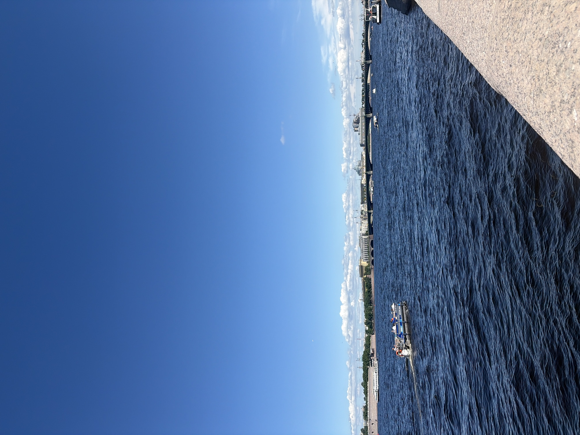 This detailed image depicts a wide river in Saint Petersburg, Russia, under a bright, clear blue sky with minimal cloud cover. The photograph is taken from a stone embankment, visible in the lower right foreground.
The sky, occupying the upper two-thirds of the frame, is a vibrant, cloudless blue, save for a thin band of small white cumulus clouds hugging the distant cityscape horizon and a single, isolated wispy cloud higher up. A faint white speck, possibly a distant aircraft, is discernible in the upper-middle section of the sky.
The Neva River dominates the mid-ground and lower-left, displaying deep blue, rippled water surfaces indicating movement. A small, open motorboat, identified by "WWW.NORD-TRAVEL.RU" on its hull, travels from left to right in the mid-left, leaving a distinct white wake. Approximately five to six passengers are visible seated within this boat. Multiple other smaller vessels are scattered across the river, and a longer white passenger boat is either docked or moving slowly near the far left bank. Another large white boat is partially visible, docked near the right bank.
The distant far bank features a dense cityscape characteristic of Saint Petersburg, including a stone embankment, historic buildings with diverse architectural styles, some featuring green domes or roofs, and areas of green foliage. A multi-span bridge, possibly a drawbridge, extends across the river, connecting the distant banks. Structures resembling fortifications or large historical complexes are visible on the far left bank.
The immediate foreground on the right consists of a light-colored, rough-textured granite or stone embankment, angled diagonally, indicating the vantage point is from a riverside promenade or quay.
