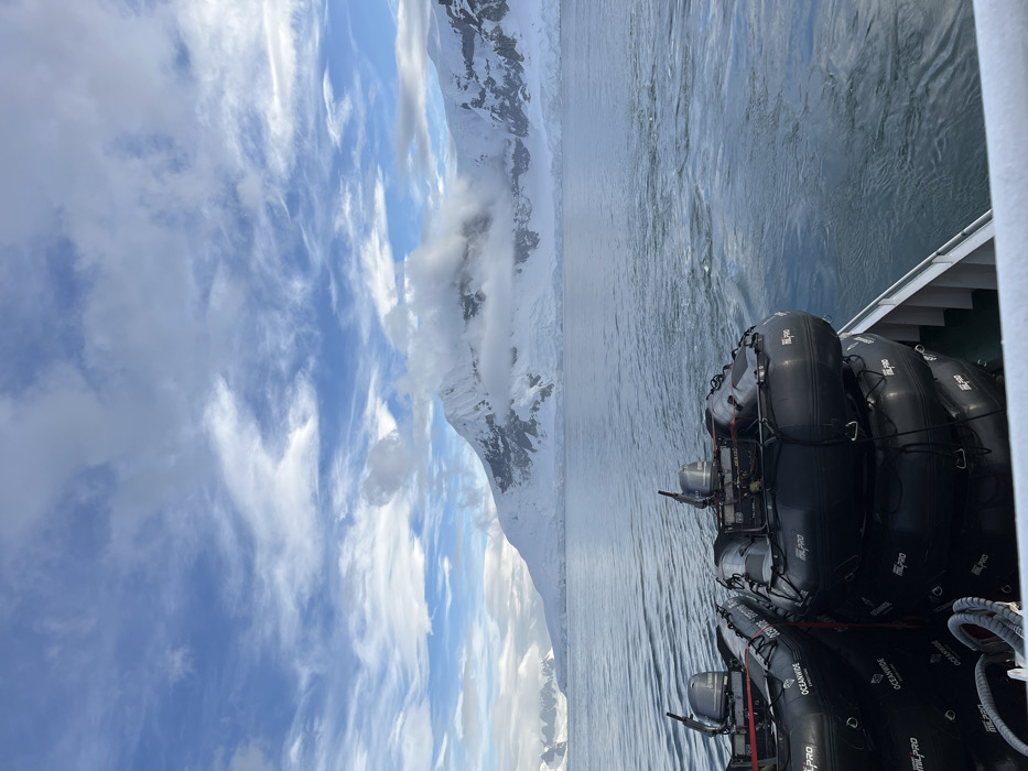 A tilted view from a boat shows calm water and a mountainous landscape in the distance. The sky is filled with scattered white clouds. On the boat's deck, several inflatable boats are stacked, taking up a significant portion of the foreground. The boats are dark-colored and appear to be ready for use.