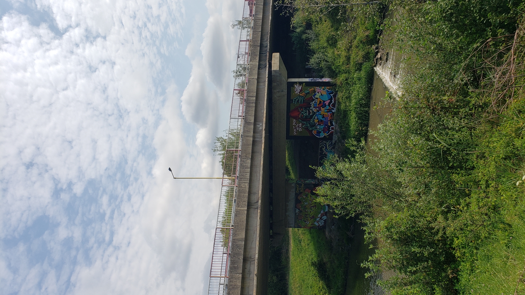 The image shows a concrete bridge spanning a body of water, likely a river or stream. The bridge has a red and white railing along its top. The underside of the bridge is adorned with colorful graffiti, including a large mural of a monstrous figure with the text "DON'T DEAL WITH DEVIL". A yellow lamppost stands on the side of the bridge, its light pointing upwards. Lush green grass and dense foliage, including trees and bushes, surround the water and the bridge's supports. The sky is partly cloudy with scattered white clouds against a light blue background. The scene appears to be an outdoor setting, possibly in a park or along a waterway. The location is identified as being in the city of Prešov, Slovakia.