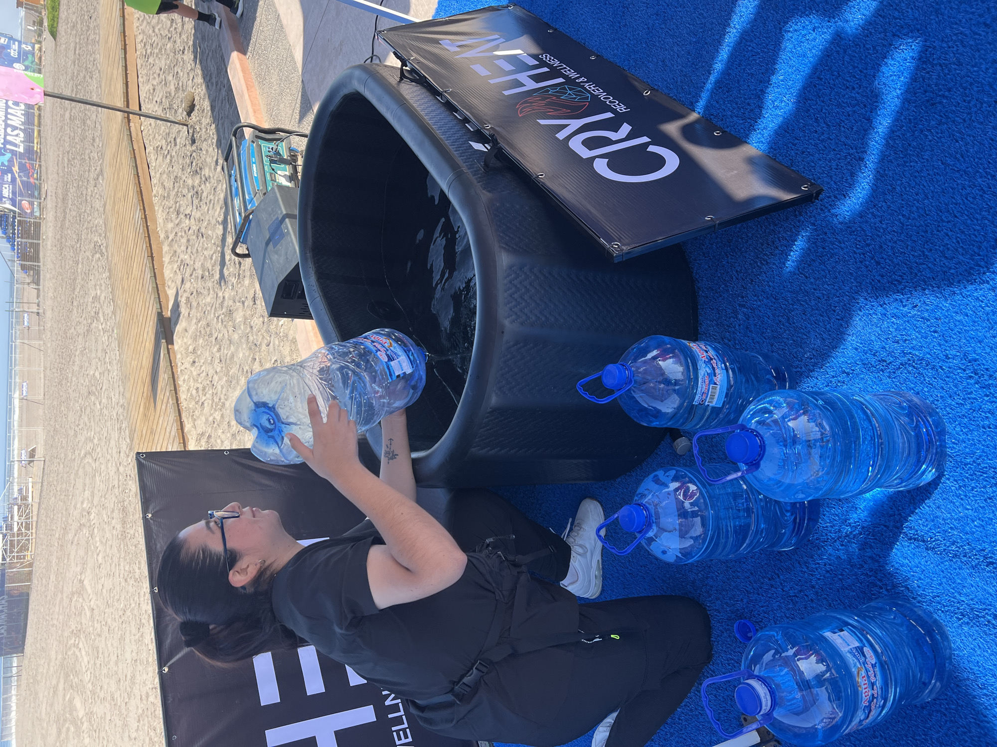A medium shot of an individual in dark clothing adding water to a black tub, with several large water bottles nearby. The individual is fair-skinned with dark hair pulled back, wearing glasses, and has a tattoo on their forearm. They are kneeling on a blue, carpet-like surface. Adjacent to the tub is a sign that reads "CRY/HEAT RECOVERY & WELLNESS". The background shows a beach scene with a wooden boardwalk, sand, and stadium seating in the distance. A sign that states LAS MAC further indicates the image being located within the city of Arica, Chile.