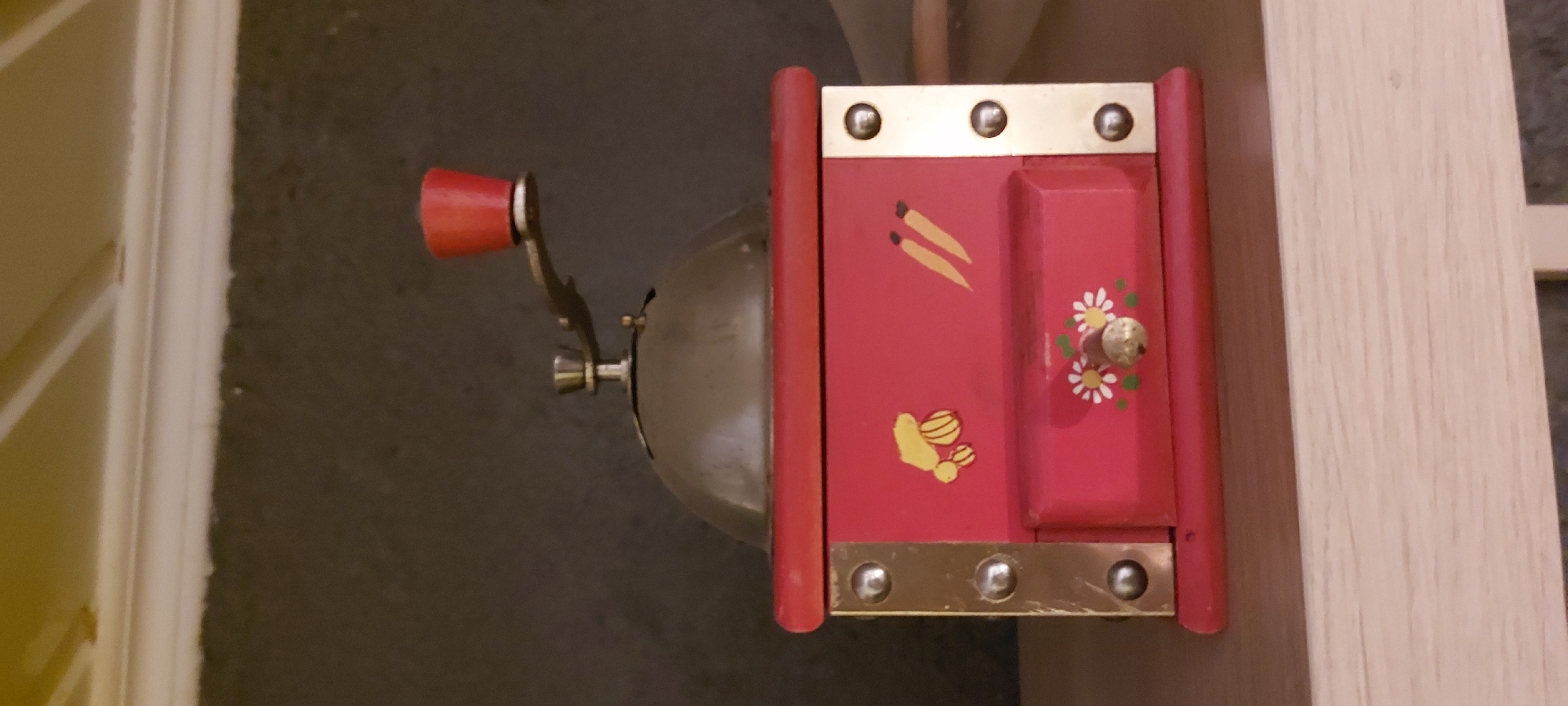 A red, wooden coffee grinder with a metal top and a crank handle with a red knob sits on a wooden shelf. The body of the grinder is decorated with painted images of bananas on the left and carrots on the right. A small drawer with a floral design and a brass knob is located on the lower part of the grinder. The background is a dark, textured wall. The overall scene appears to be an indoor setting, likely a home. There is no identifiable location in the city of Riga, Latvia.