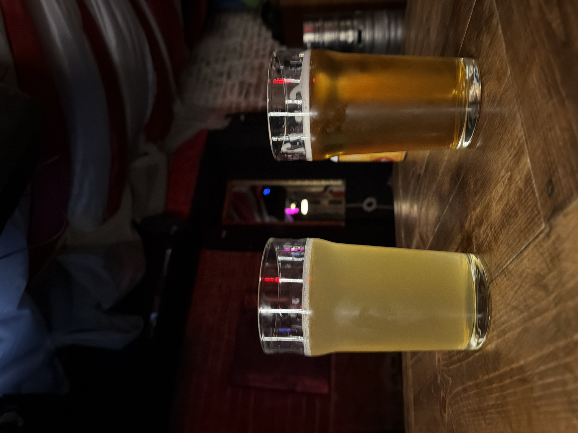 The image shows two glasses of beer on a wooden surface. The top glass contains a darker amber beer, while the bottom glass contains a lighter, more opaque beer. Behind the glasses, there is a blurred background with hints of furniture and what appears to be a red, white, and blue patterned fabric. Reflections of lights are visible in the glass. There is not enough information to determine if this picture was taken in Rostov-on-Don, Russia.