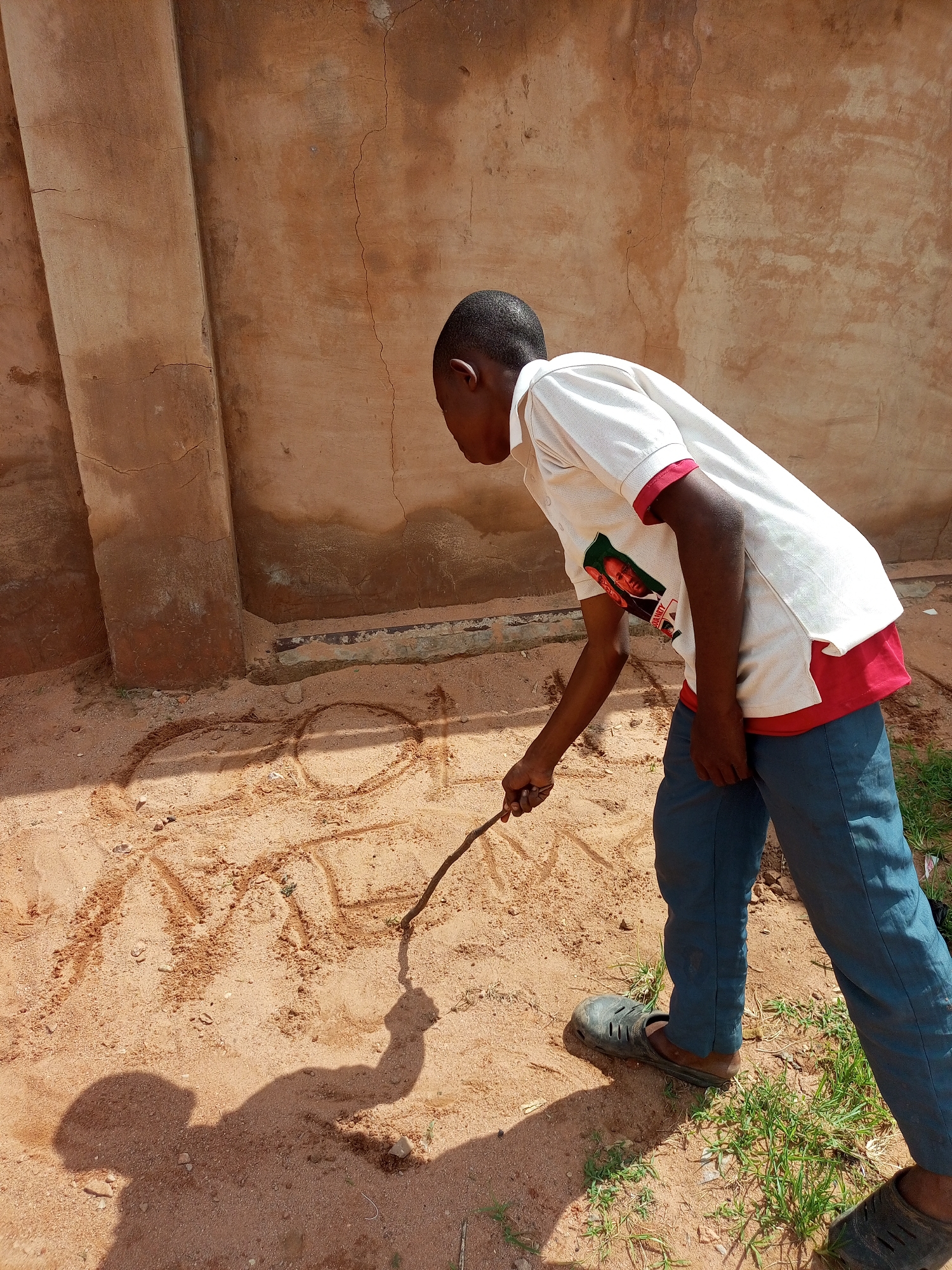 A young boy, likely under 10 years old, is drawing in the dirt with a stick. He is wearing a white polo shirt with a red collar and trim, and dark blue pants. The shirt has a graphic of a person's face on it. He is wearing dark, slip-on shoes. The ground is covered in reddish-brown dirt and sand, with some patches of green grass. Behind him is a textured, beige-colored wall with vertical, recessed panels. The scene appears to be outdoors, possibly in a courtyard or an open area next to a building. The sun is casting a shadow of the boy and his stick onto the ground. The overall environment suggests a warm, dry climate. The drawing in the dirt is rudimentary, consisting of loops and lines. The location is described as the city of Tempure, Nigeria.