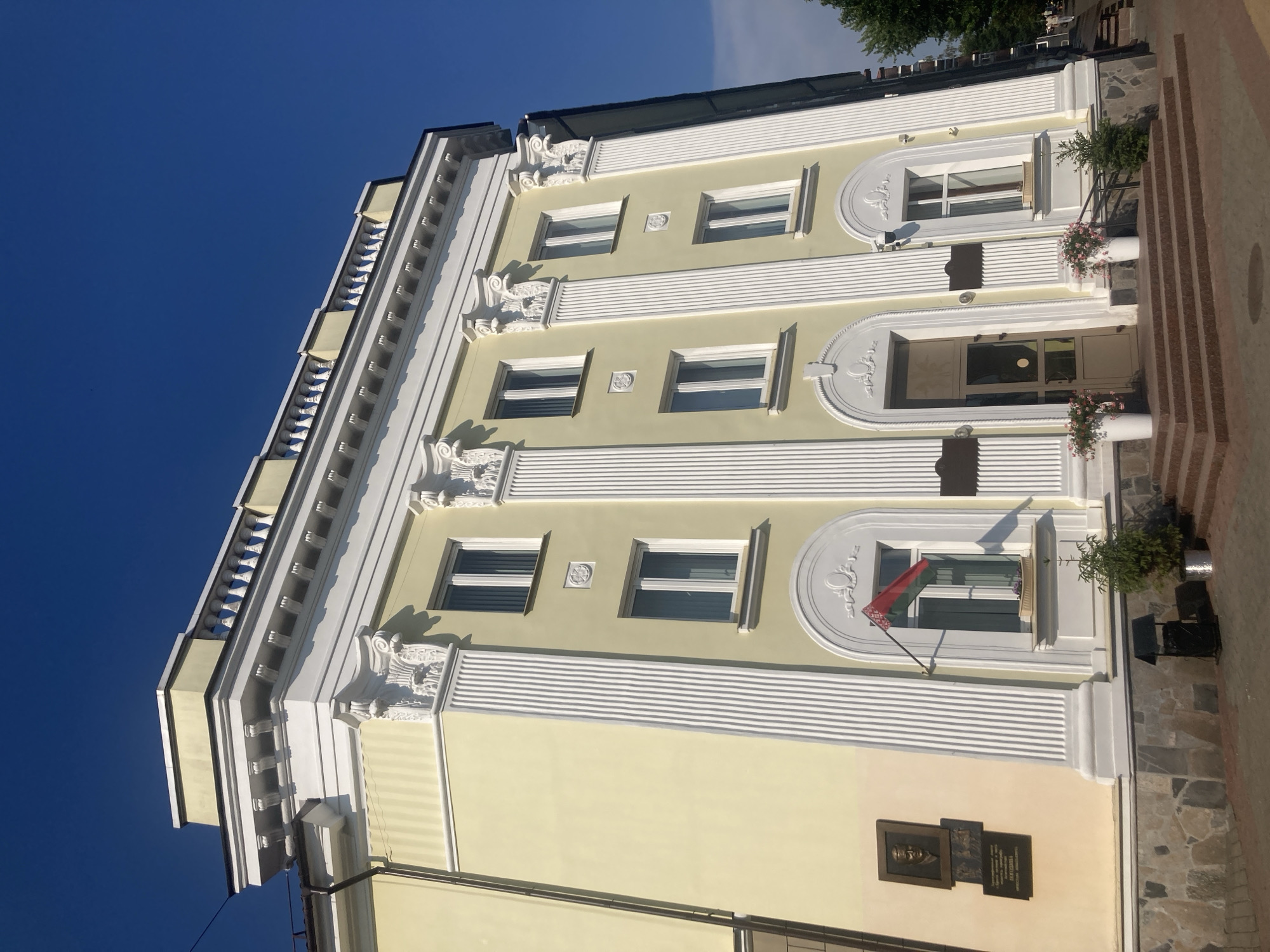 The image shows the facade of a pale yellow building, likely a government or public institution, located in Brest, Belarus. The building features classical architectural elements such as fluted pilasters, ornate moldings, and a decorative balustrade along the roofline. There are several windows on the upper floors, and a main entrance with a glass door is visible on the ground floor, approached by a set of granite steps. A flag of Belarus is displayed from one of the windows, and a bronze plaque with a portrait and text is mounted on the wall to the left of the entrance. Two planters with pink flowers flank the entrance, and a small evergreen tree is situated near the steps. The sky is clear and blue, indicating a sunny day. The scene is quiet and orderly, with no people prominently featured in the immediate foreground.