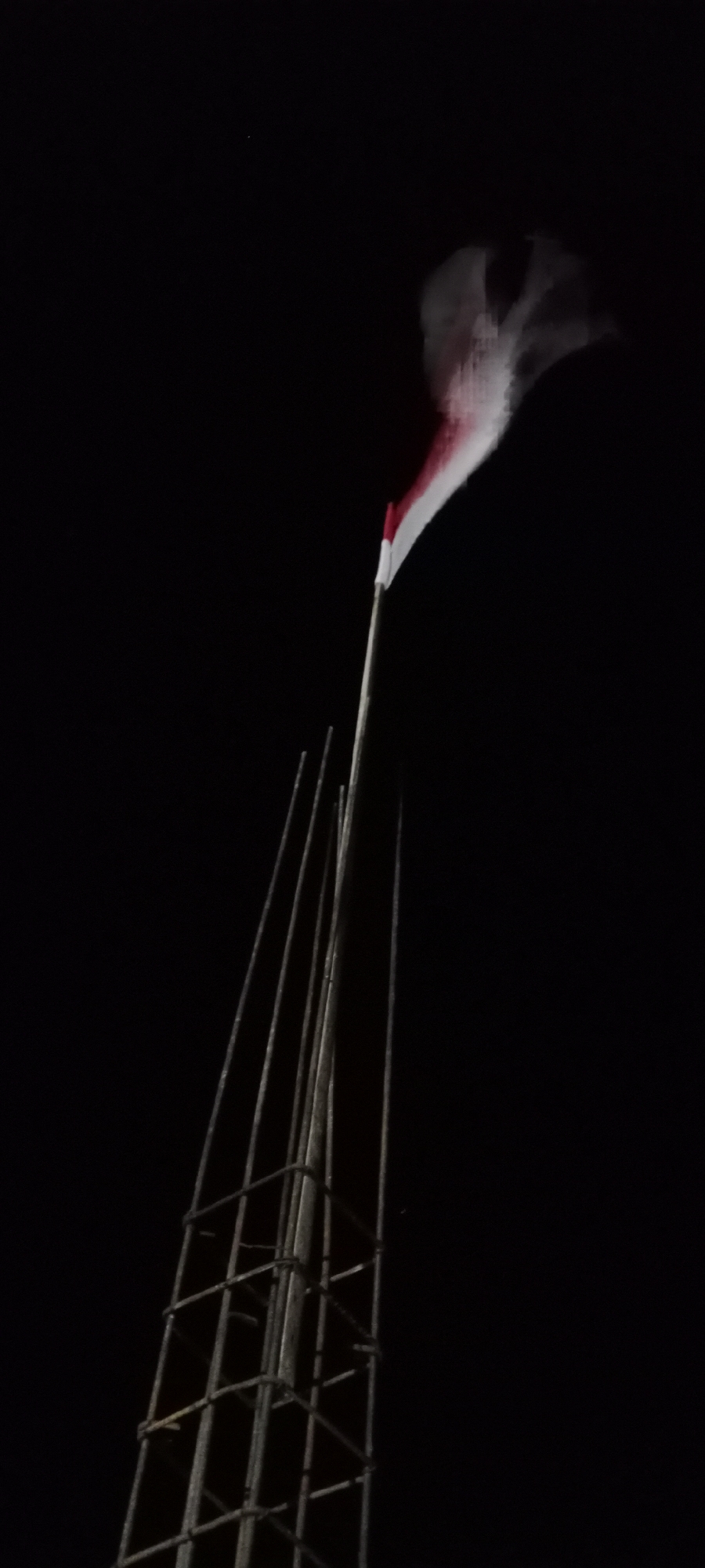 The image shows an Indonesian flag waving against a black night sky. The flag has two horizontal bands of color: red on top and white on the bottom. The flag is attached to a tall pole made of bamboo or similar material, which extends upwards from a partially constructed structure of rebar. This structure appears to be a foundation or pillar for a building. The scene is dark, suggesting it is nighttime. There are no people visible in the image. The location is identified as Soko, Indonesia.