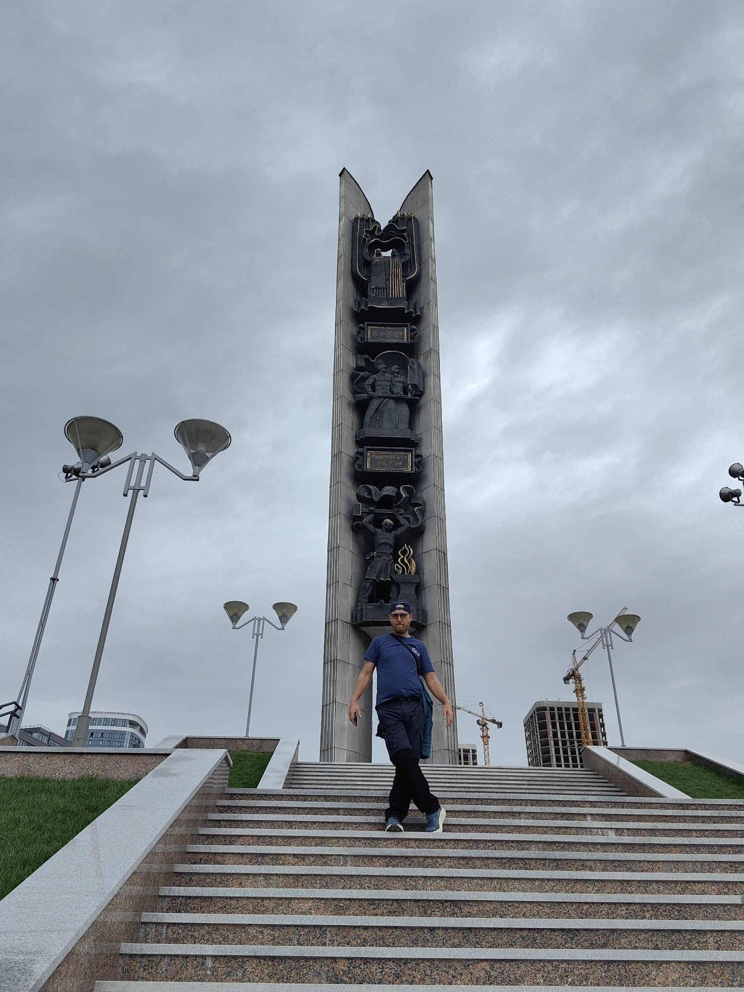 A medium shot captures a man standing on a wide set of steps leading up to a tall, Soviet-era monument. The monument is a dark grey color and features relief carvings depicting figures and symbols. A building and two construction cranes are visible in the background, partially obscured by the monument. The sky is overcast. Several street lamps are visible on the sides of the stairs. The man is wearing a blue shirt, dark pants, and a baseball cap, and is standing with his legs crossed and arms outstretched. The location is Izhevsk, Russia.
