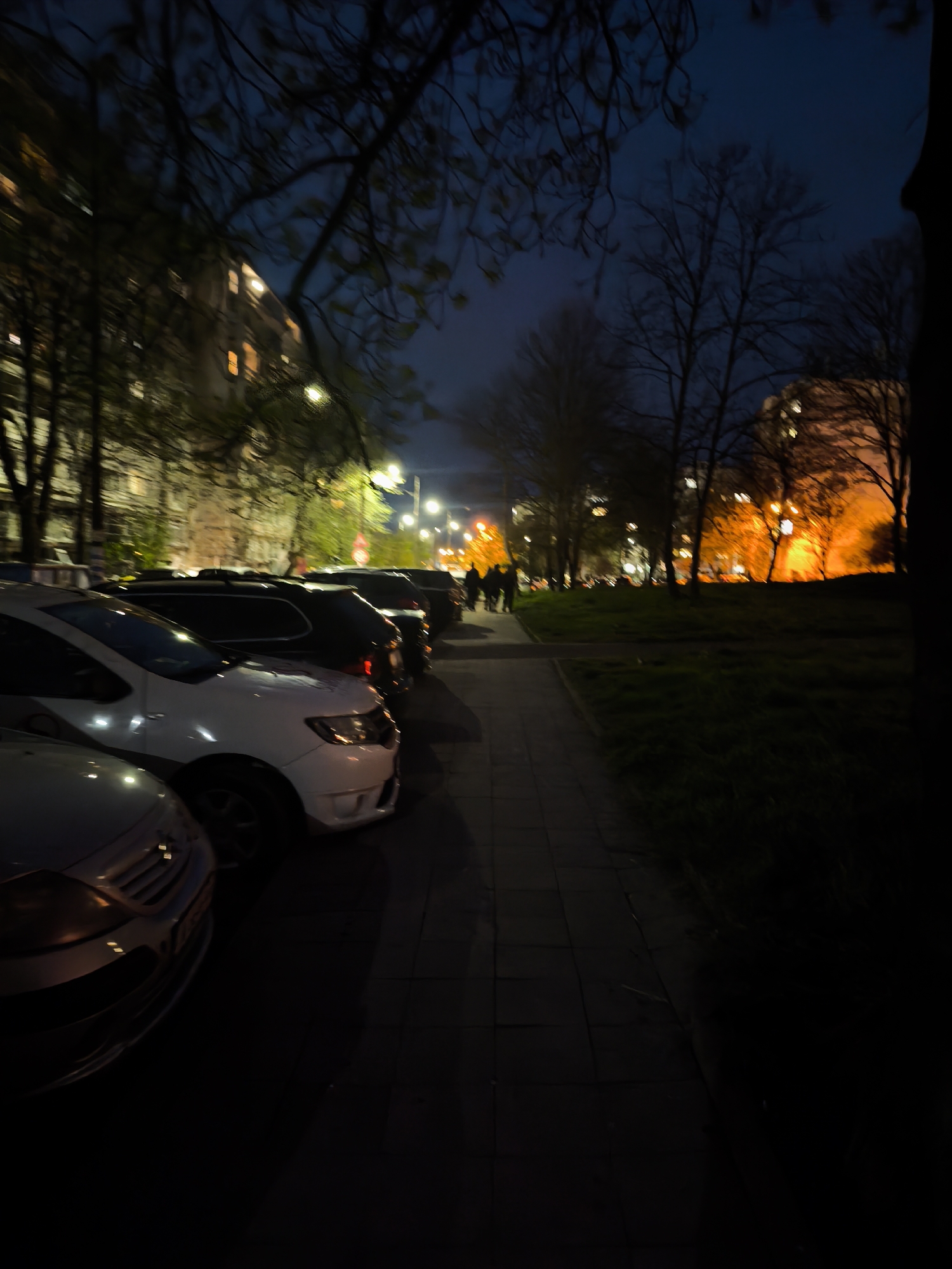 The image depicts an urban street scene at night or late evening in Burgas, Bulgaria. On the left, a line of parked cars is visible, starting with a silver/grey vehicle in the foreground, followed by a white car, and several darker vehicles further down the street. A paved sidewalk runs alongside the cars, extending into the distance.
Behind the parked cars and trees, multi-story buildings with illuminated windows can be seen, hinting at residential or commercial spaces. On the right side of the sidewalk, a grassy area, possibly a park or green strip, is partially visible, flanked by more trees. Some trees appear bare, while others retain sparse foliage, suggesting late autumn or early spring.
In the mid-ground, a group of three to five indistinct figures are walking on the sidewalk, illuminated by bright streetlights. The background features additional buildings and a prominent orange glow, likely from artificial lighting, casting a warm hue on distant trees and structures. The sky is a dark blue-black, indicative of nighttime. Artificial lights from streetlamps and building windows create pockets of brightness against the overall darkness. The image has a slightly blurry, atmospheric quality, common in low-light conditions. No discernible text is visible.