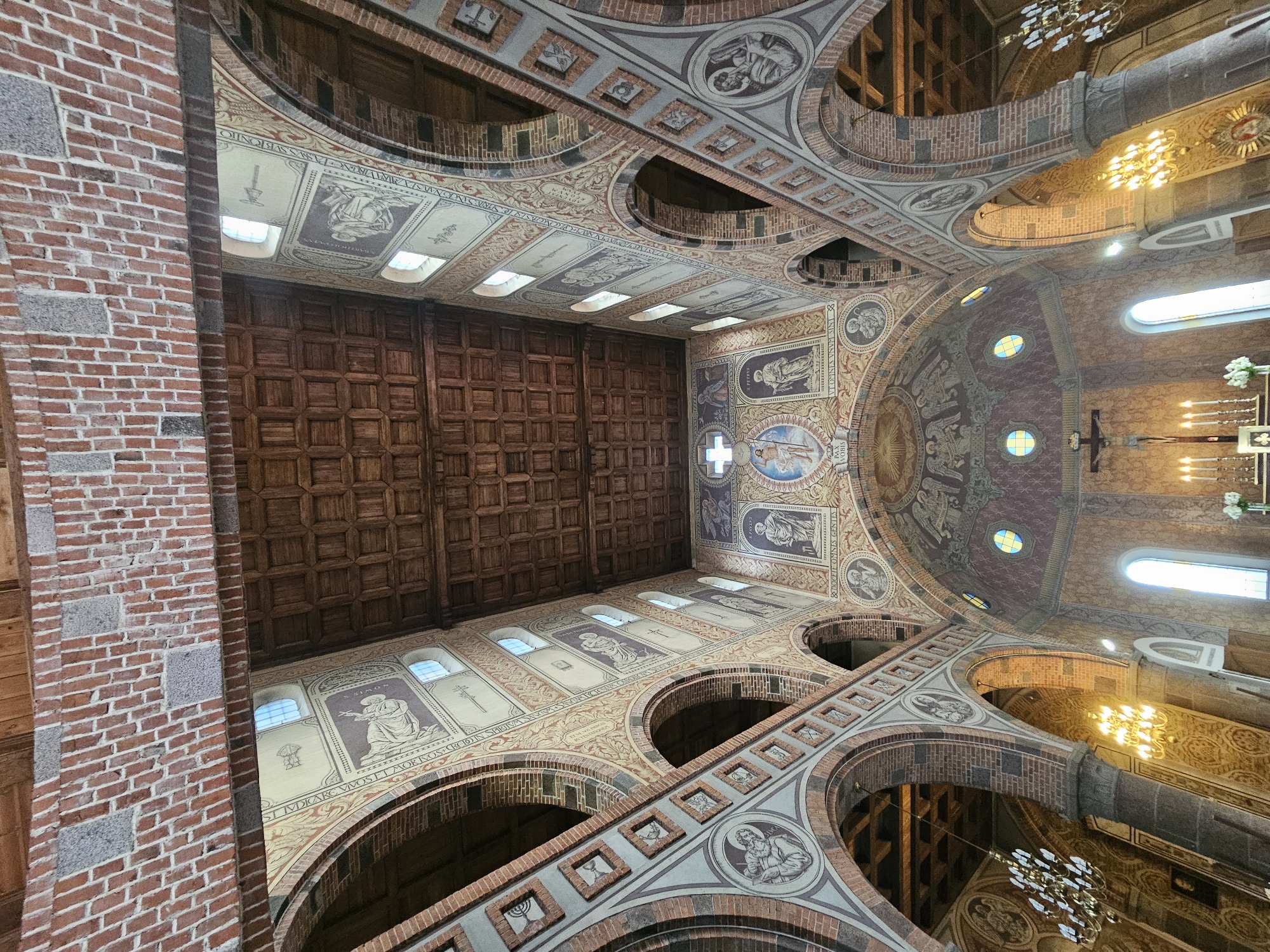 The image shows the interior of the Church of the Assumption of the Blessed Virgin Mary in Lentvaris, Lithuania. The photograph is taken from a low angle, looking up at the ceiling and the architecture of the church.
The ceiling is made of dark wooden planks, arranged in a coffered pattern. Supporting the ceiling are massive brick arches and columns. The walls are adorned with numerous fresco paintings, depicting biblical scenes and figures. These frescoes are in muted tones of beige, brown, and red, with some highlights of blue and yellow in the windows.
Along the nave, on the upper walls, are a series of arched windows letting in natural light. Below the windows, the walls are decorated with bands of intricate patterns and framed artwork, likely further religious imagery. In the foreground, pillars with decorative stone bases are visible, leading the eye towards the altar area.
The altar itself is not fully visible, but a cross with a figure of Christ is prominent, flanked by several lit candles and a small bouquet of white flowers. Chandeliers hang from the ceiling, providing artificial light, and some have an ornate, multi-tiered design. The overall impression is one of grandeur and historical significance, characteristic of old European churches. The architecture suggests a Romanesque or Gothic influence with the arched openings and stone construction.