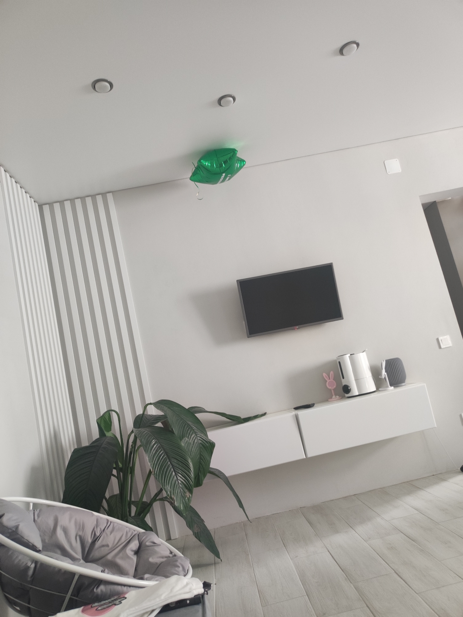 The image shows an interior space, likely a living room, featuring a white wall with vertical striped design on the left side. A green balloon is stuck to the ceiling. A wall-mounted television is present, positioned above a white, floating cabinet. The cabinet holds a pink rabbit figurine, a humidifier, and a gray object. In the lower-left corner, a baby swing or bassinet is visible with gray fabric. A large green plant is located next to the swing. The floor is covered with light-colored wood-look planks. The location within the city of Votkinsk, Russia is not possible to determine from the image.