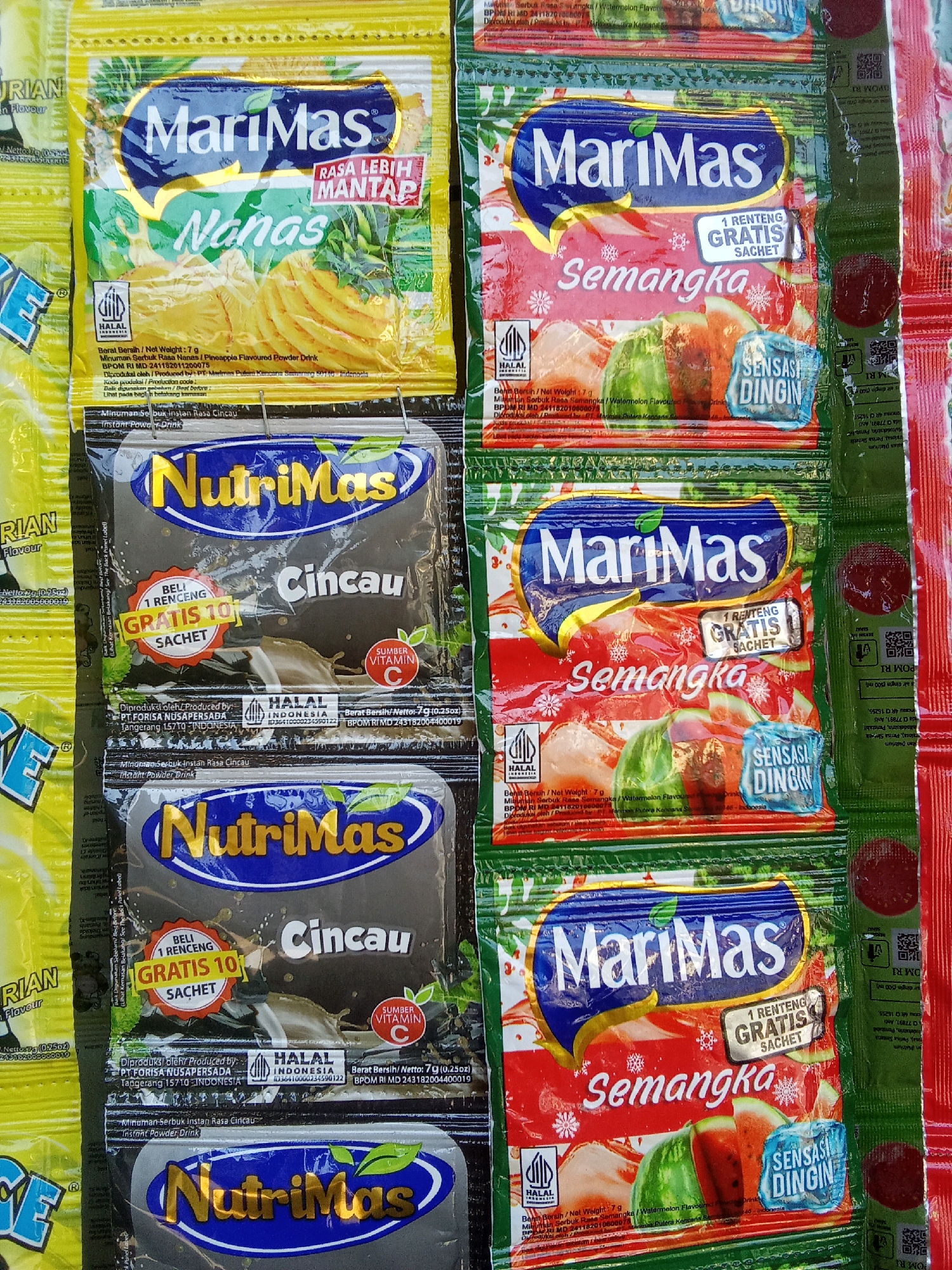 The image displays a vertical arrangement of several packaged drink mixes, likely in a market or store setting in Sumpiuh, Indonesia. The products are predominantly from two brands: "MariMas" and "NutriMas."
The "MariMas" packages are colorful, featuring images of fruit. One type, in a red and green wrapper, advertises "Semangka" (Watermelon) flavor with the text "SENSASI DINGIN" (Cold Sensation) and "1 RENTENG GRATIS SACHET" (1 String Free Sachet). Another "MariMas" package, yellow, is labeled "Nanas" (Pineapple) and mentions "RASA LEBIH MANTAP" (More Delicious Taste).
The "NutriMas" packages are darker, with gold and white lettering. One prominent "NutriMas" package is for "Cincau" (Grass Jelly) flavor, featuring "BELI 1 RENCENG GRATIS 10 SACHET" (Buy 1 String Get 10 Sachets Free) and "SUMBER VITAMIN C" (Source of Vitamin C). Another "NutriMas" package below it also appears to be for "Cincau" flavor.
All packages bear "HALAL" certification logos. Text on the packaging is in Indonesian and English, indicating net weight (7g or 7g (0.25oz)) and manufacturer details, such as "PT. FORISA NUSAPERSADA Tangerang 15710, INDONESIA." The items are stacked closely together, suggesting they are for sale. The overall lighting is bright, and the focus is on the products themselves.