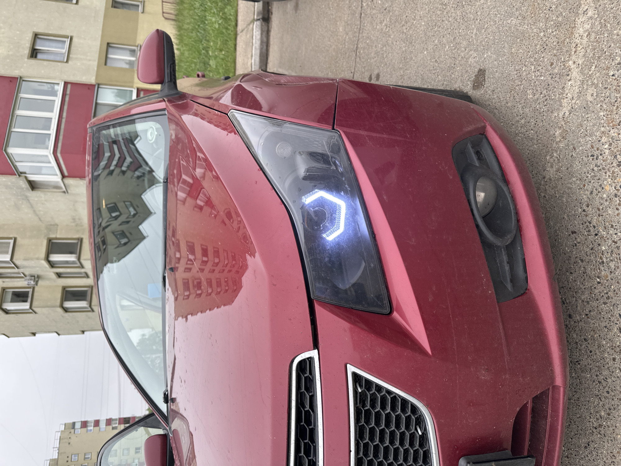 A reddish-burgundy car is parked outdoors, viewed from an angled perspective that tilts the image. The front-right side of the car is prominent, featuring a custom headlight with a bright, hexagonal LED daytime running light. The car's front grille, partially visible, has a honeycomb pattern with silver trim. The bumper includes a housing for a fog light, which appears to be missing or deeply recessed. The car's body panels show some dust and minor scuffs, and the paint has a metallic sheen.
Reflections of a multi-story residential building with numerous windows and balconies are visible on the car's windshield and hood. In the background, another cream-colored building with red sections and many windows stands against an overcast sky. Patches of green grass are seen near the buildings. The car is situated on a textured concrete surface.
The weather appears to be cloudy, and the car and ground show subtle signs of dampness or dust, suggesting a recent rain or general outdoor exposure on a drab day. There are no people or discernible text visible in the image. The architectural style of the buildings suggests an urban or suburban environment, consistent with locations in Chaikovsky, Russia.