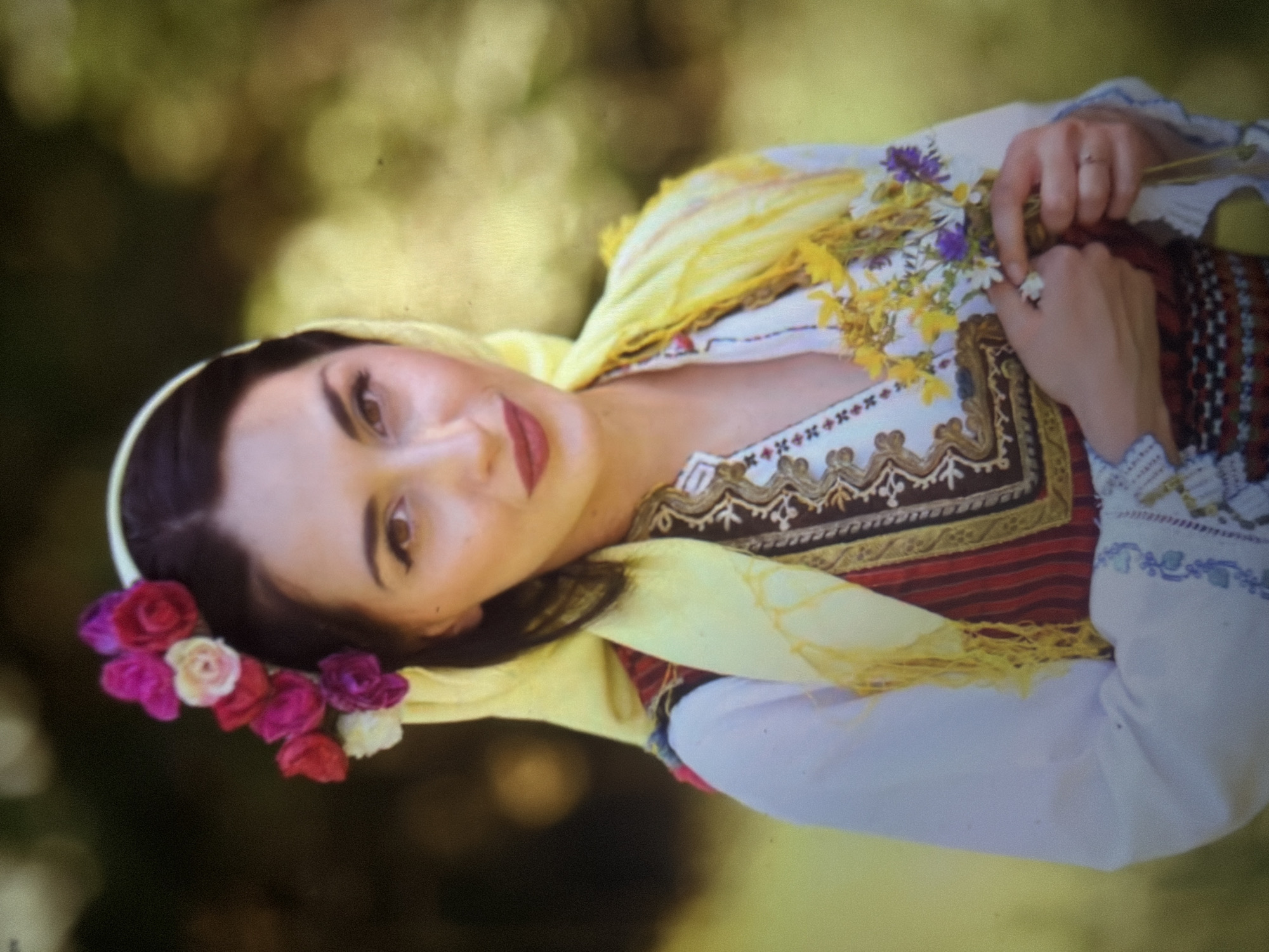 A young woman, approximately in her late teens to early twenties, is depicted in a half-length portrait, standing outdoors in Shumen, Bulgaria, against a heavily blurred green and yellow-green natural background of foliage.
She has dark brown hair, parted centrally, tucked beneath a light yellow headscarf that drapes over her shoulders. A cluster of artificial fabric flowers in shades of fuchsia, red, cream, and deep purple adorns the left side of her head (viewer's left) atop the headscarf. Her face features brown eyes with visible winged eyeliner, defined eyebrows, and lips colored with a dark reddish-brown lipstick. She presents a gentle smile and looks directly forward, engaging with the camera.
She is dressed in traditional Bulgarian folk attire. This includes a white long-sleeved blouse with intricate blue and purple embroidery on the cuffs. Over the blouse, she wears a brown vest or bodice, richly embroidered with gold, white, and brown geometric and stylized floral patterns around the neckline and front. Below the vest, a striped fabric in red and dark tones is visible, followed by a band with further dark geometric embroidery.
Her hands are clasped in front of her, holding a small, loose bouquet of wildflowers. The bouquet consists primarily of yellow, white (daisy-like), and purple/blue blossoms with green stems. A thin, plain silver-colored ring is visible on the ring finger of her right hand.
The outdoor environment is characterized by soft, natural lighting. The background blur indicates a shallow depth of field, isolating the subject. There are no other visible people or objects in the scene.