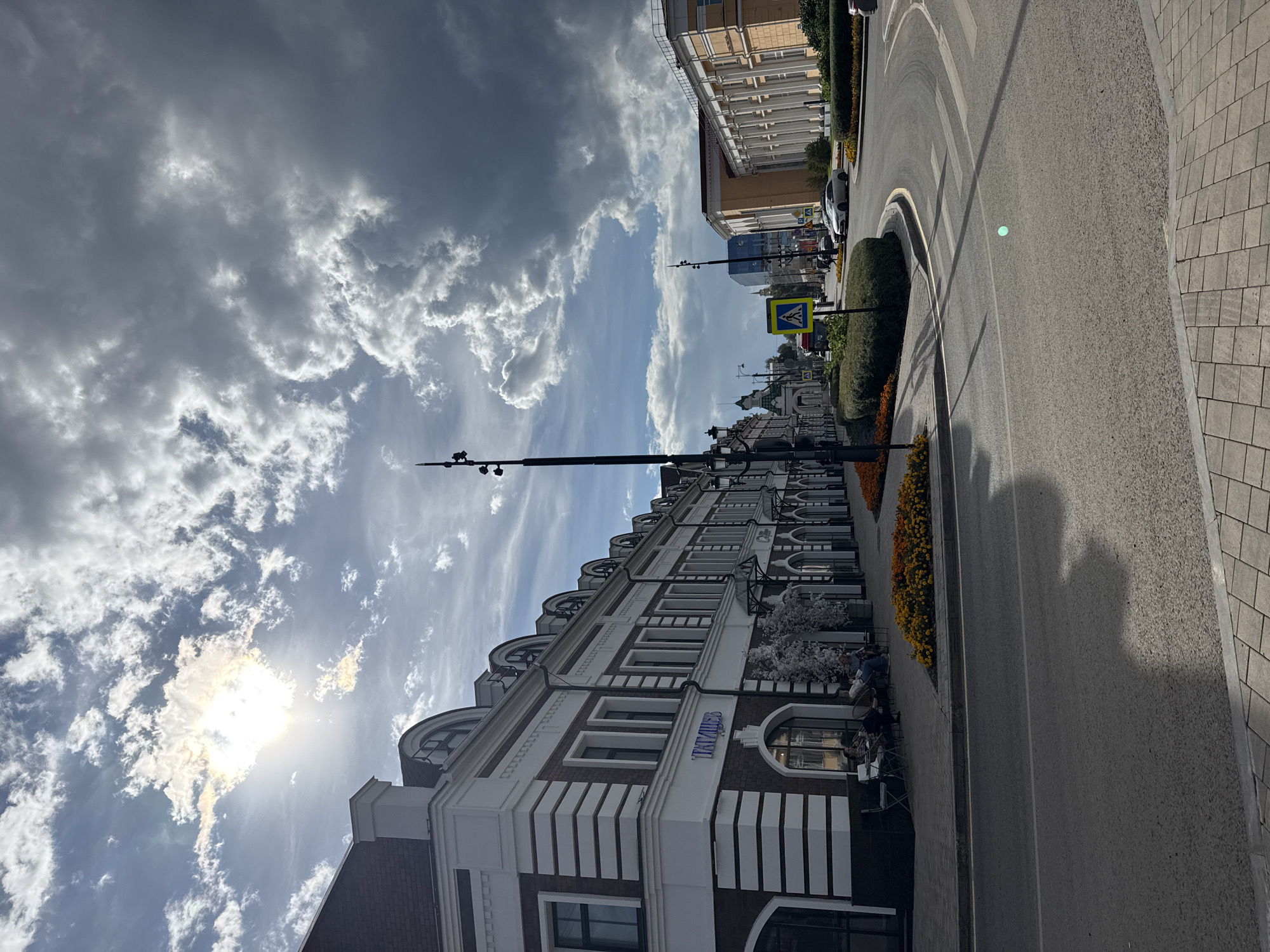 The image depicts a street scene in the city of Perm, Russia, likely on a street with a mix of historic and modern architecture. The sun is shining brightly, partially obscured by dramatic, dark storm clouds, creating a contrast between light and shadow.
In the foreground, a paved road curves to the right, with a pedestrian crossing marked by white stripes. The pavement is made of cobblestones. A shadow of a person or object is cast across the road.
To the left, a row of buildings features a classic facade with white trim and dark brickwork. Some of these buildings have arched windows and decorative elements. People are seated at tables outside one of the buildings, possibly at a cafe. A blue sign with Cyrillic lettering is visible on this building.
Further down the street, more buildings are visible, including a large, light-colored building with columns on the right side of the image. Traffic lights and street lamps are also present. A yellow pedestrian crossing sign is posted on a pole. A white car is visible on the road.
The sky is a prominent feature, with a mix of bright blue sky and heavy, gray clouds. The overall impression is one of a dynamic weather day in an urban environment.