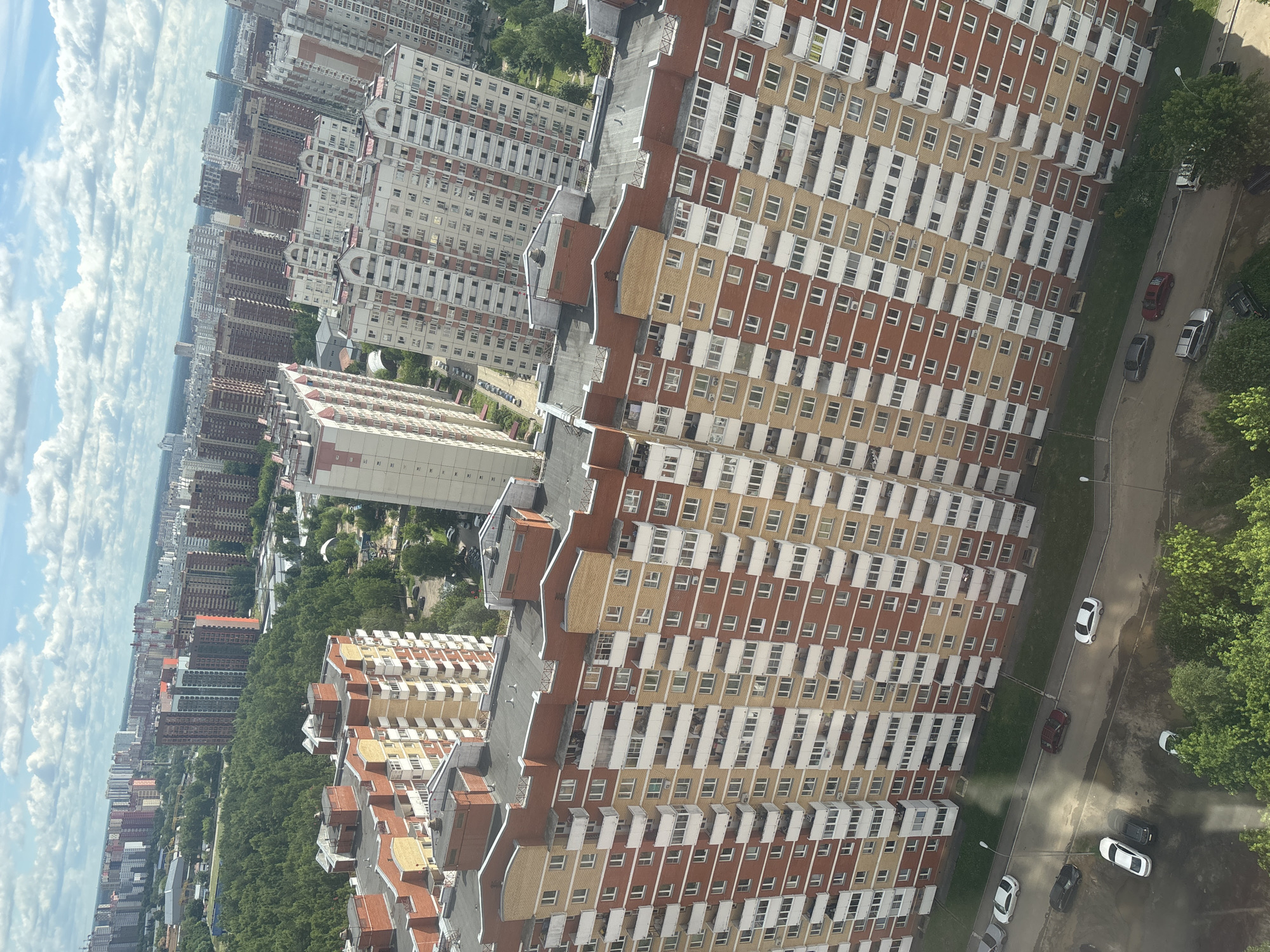 This is a high-angle aerial view of a dense urban residential area in Гознаковец Снт, Russia. The foreground is dominated by a massive, multi-story apartment building with a red brick and cream-colored facade, featuring numerous balconies and windows. The building's roof is flat with some mechanical structures. To the left and behind this building, other similar apartment complexes of varying heights and architectural styles extend into the distance, creating a sprawling cityscape.
Below the apartment buildings, a paved road winds through the scene. Several cars are parked on or near the road, including a white sedan, a dark SUV, a dark-colored car, and a red sedan. Green trees and some patches of grass are visible interspersed between the buildings and along the roadside, indicating a suburban or semi-urban setting within the city. The sky is partly cloudy with white cumulus clouds against a blue backdrop. The overall impression is one of a large, modern housing development.