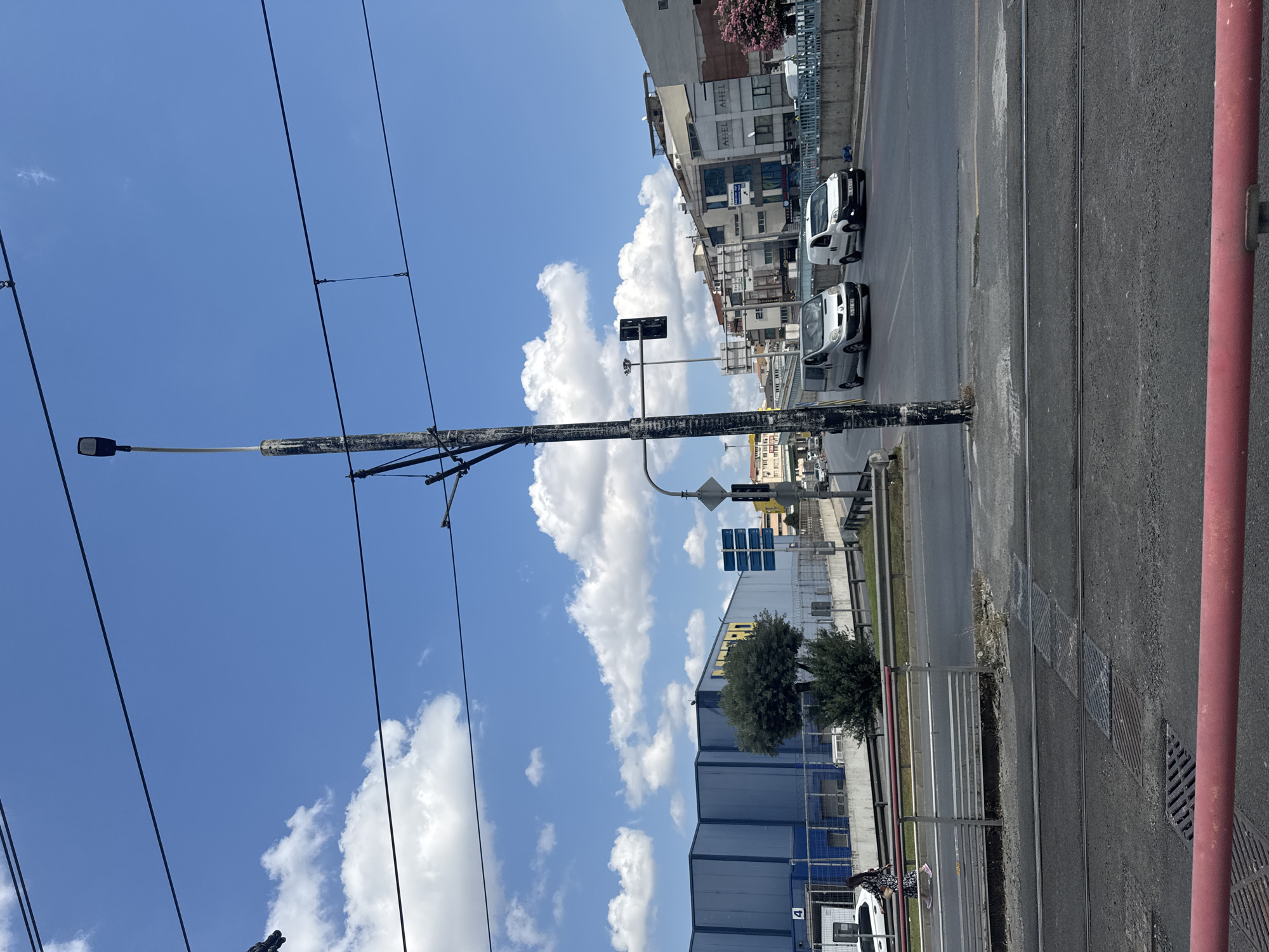 The image shows a street scene in Bahçelievler, Turkey, on a sunny day with a blue sky and scattered white clouds. In the foreground, there are tram tracks on an asphalt road. A woman in a patterned dress and pink shoes is walking on a sidewalk to the left of the road, near a blue building. A white car is parked to her left.
Further down the road, several vehicles are driving, including two vans, one silver and one white. A traffic light, showing a red light, is visible on a pole. In the background, there are multi-story buildings with shops and apartments, and some trees. A large blue building with yellow lettering that reads "METRO" is prominent on the left side of the image. Overhead, power lines and a street lamp are visible.