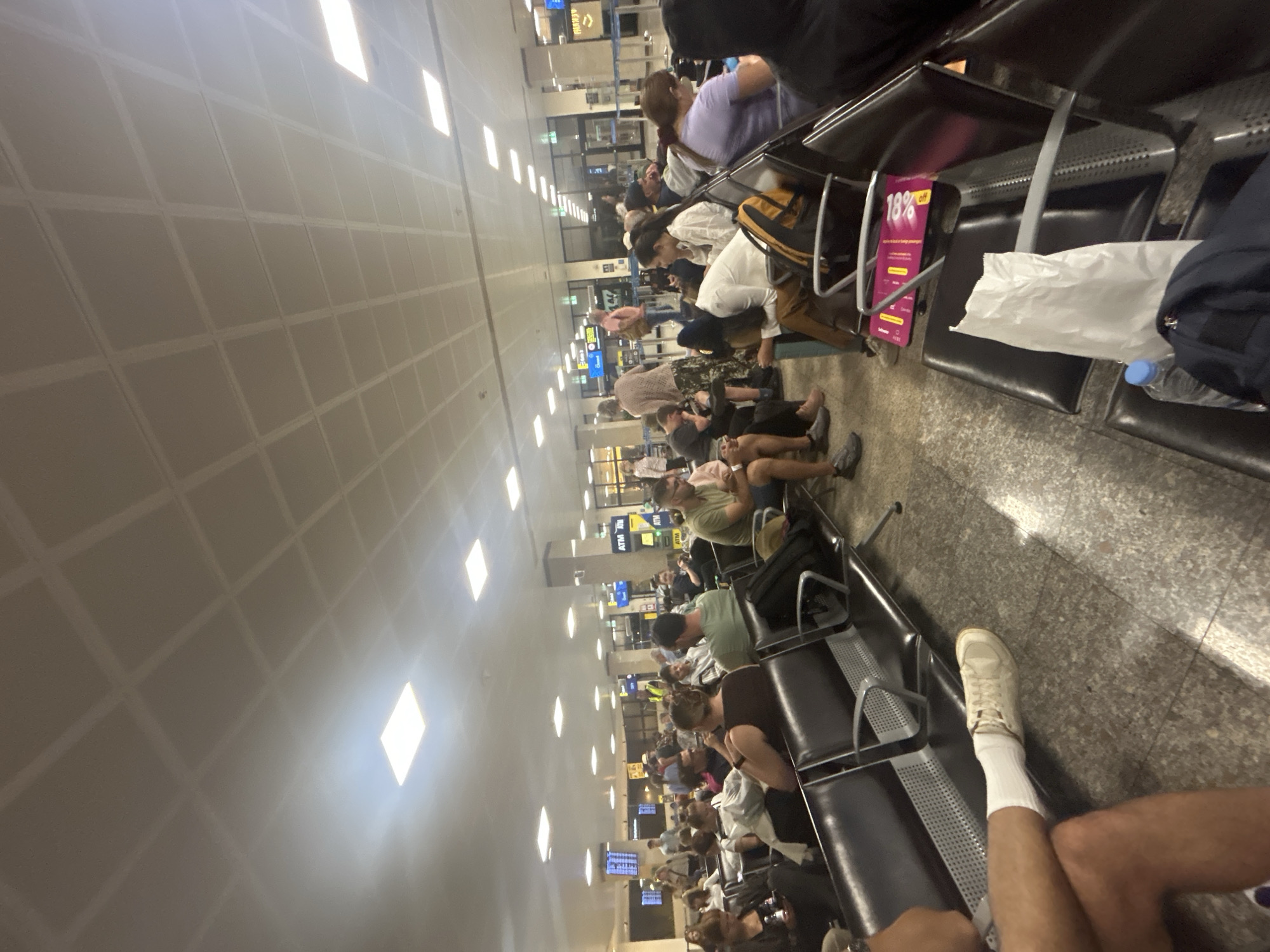 The image shows a crowded waiting area at Malta International Airport in Luqa, Malta. Passengers are seated on rows of black, airport-style seating, waiting for their flights. The scene is brightly lit by overhead fluorescent lights recessed into a white, grid-patterned ceiling. People of various ages and ethnicities are present, some looking at their phones, others resting. Luggage, including backpacks and bags, is placed on the floor or on the seats next to the passengers. In the foreground, a person's legs are visible with a white sneaker and white socks. Further down the waiting area, ATM machines and arrival/departure boards are visible.