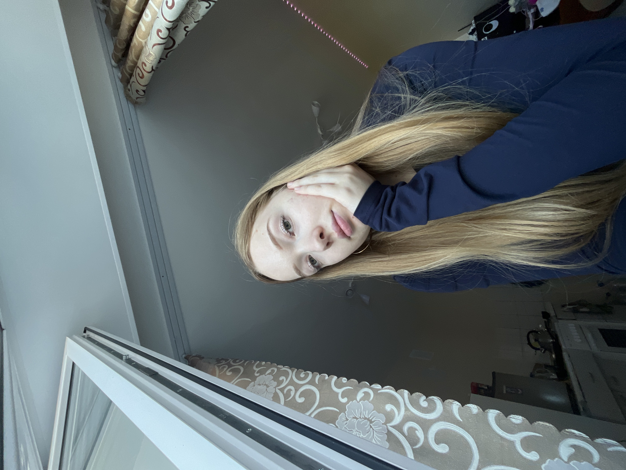 A selfie of a blonde-haired woman in Chaikovsky, Russia. She is wearing a dark blue long-sleeved shirt. Her face is partially covered by her hand. The photo is taken indoors, near a window with white frames and patterned beige curtains. A faint pink LED strip is visible on the ceiling. In the background, a desk or table is partially visible with various objects.
