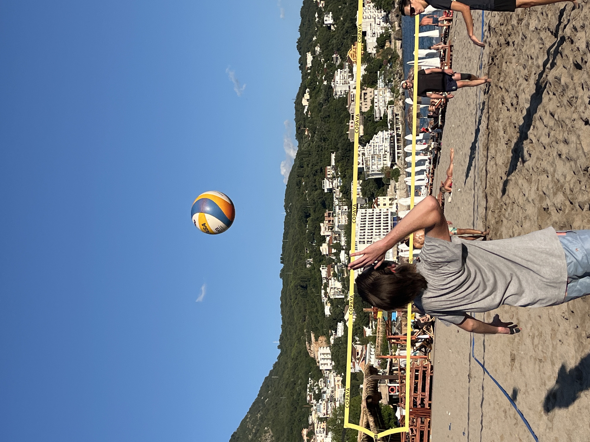 The image depicts a beach volleyball game taking place on a sunny day with a clear blue sky. In the foreground, a person with long brown hair is facing away from the camera, mid-action, with their arm raised as if to hit a volleyball, which is visible in the air above them. They are wearing a grey t-shirt and light blue shorts. The volleyball is multi-colored with yellow, blue, and white panels. The beach is sandy, with a volleyball net stretched across the middle ground. Several other people are present on the beach; some are playing or spectating volleyball, while others are sunbathing on loungers. In the background, there is a resort area with buildings, umbrellas, and palm trees, nestled against a backdrop of a lush green, mountainous hillside dotted with more buildings. The sea is visible in the far background, and the overall setting suggests a coastal resort town. The location is identified as Zankovići, Montenegro.