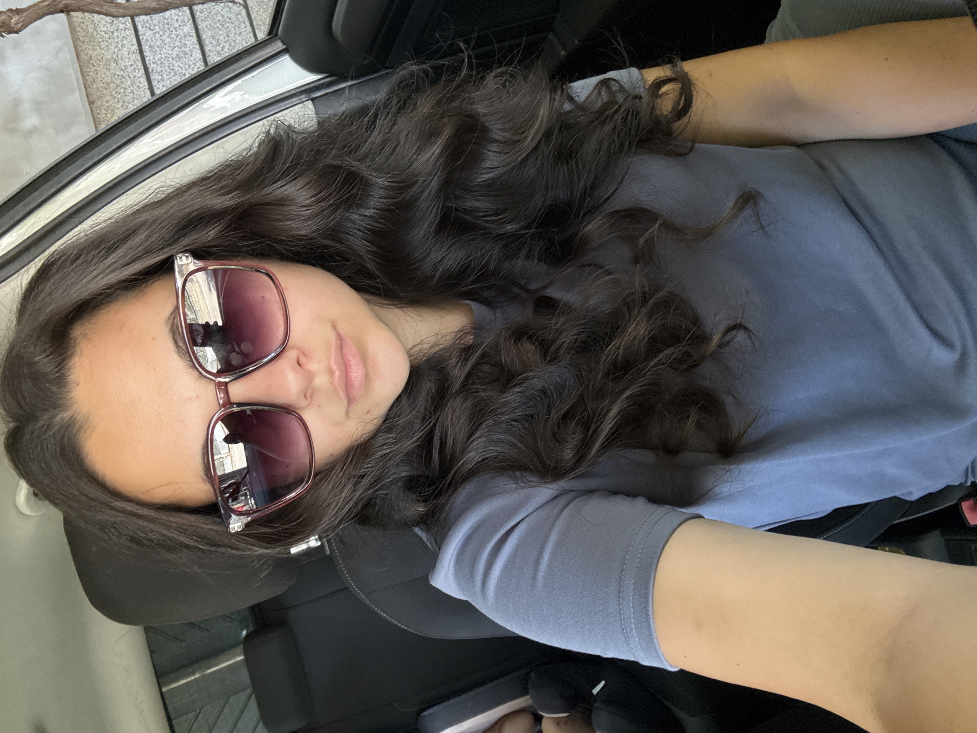 A selfie taken by a woman inside a vehicle. The woman has long, dark, wavy hair and is wearing sunglasses with a reddish-brown frame. She is wearing a grey t-shirt. The background shows the interior of the car, including a black headrest and seat. The image is taken from a slightly high angle, looking down at the woman. The location is Oreshak, Bulgaria.