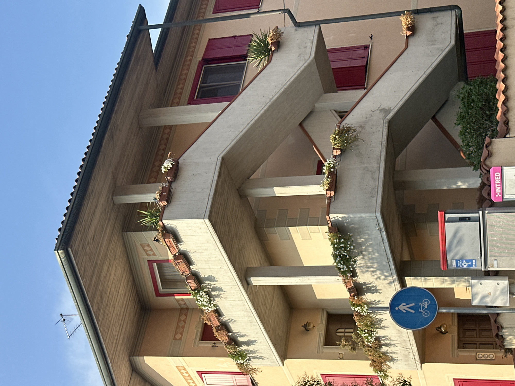This is a medium-angle outdoor shot, oriented vertically, of a building's facade with a clear blue sky above. The architecture features prominent concrete staircases that zigzag up the building, adorned with rows of planters containing white flowers and some greenery. The building itself is painted in warm, muted tones of yellow and cream, with accents of dark red for the window shutters and frames. The roof has a dark, textured tile design.
In the lower right quadrant, a blue circular sign with a white pictogram indicates a shared path for pedestrians and cyclists. Adjacent to this sign are street furniture elements, including what appears to be a mailbox or utility box and a sign with the word "INTRED" in pink.
The lighting suggests it is daytime, with bright sunlight casting distinct shadows, particularly from the planters and architectural elements onto the building's walls. The overall impression is one of a well-maintained residential or commercial building, possibly in a European town, given the architectural style and the street sign. No people are visible in the frame. The scene evokes a sense of peaceful urban life.