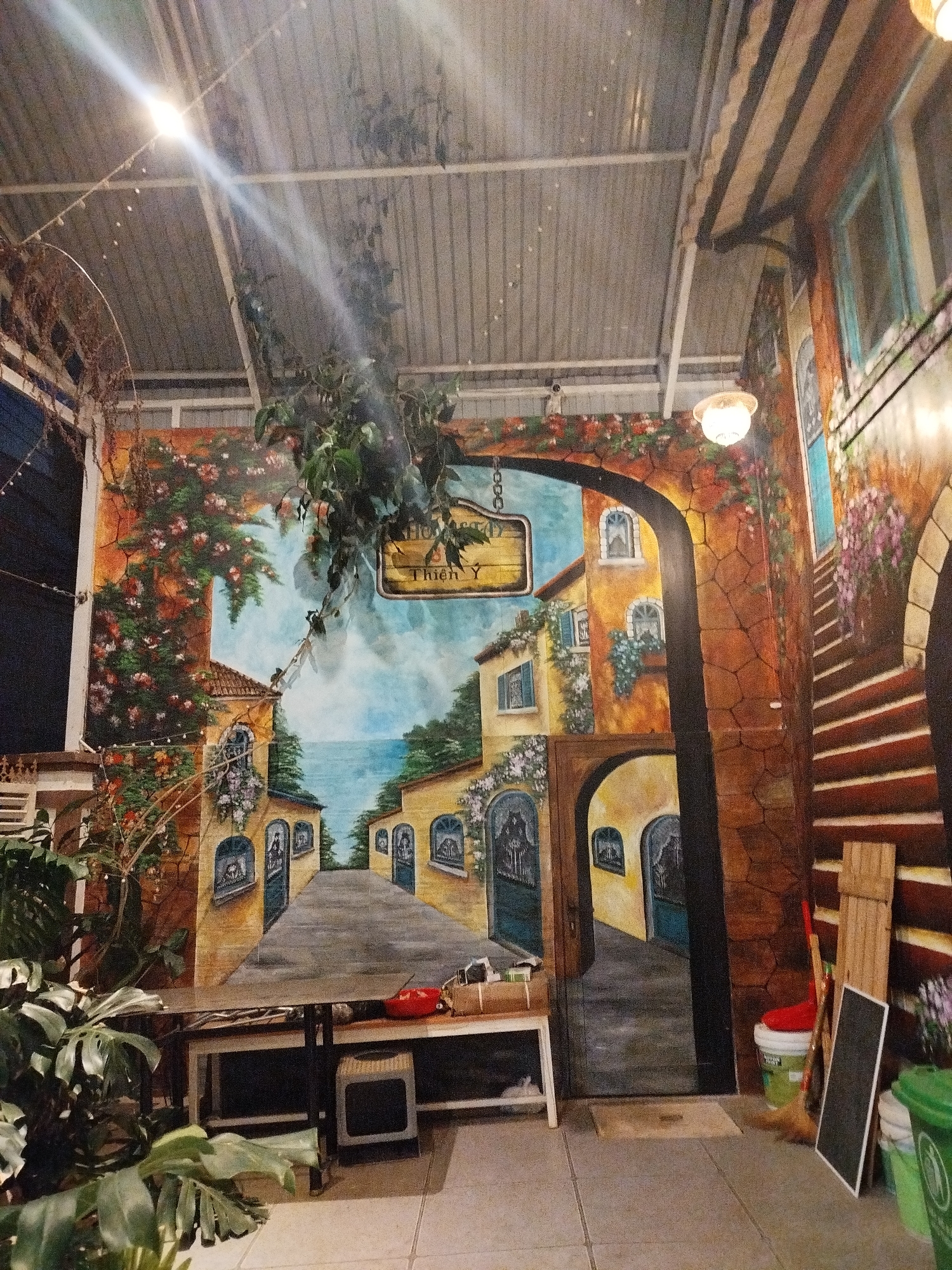 The photograph shows an indoor space in Da Lat, Vietnam, which appears to be a cafe or restaurant with a painted mural on the back wall. The mural depicts a Mediterranean-style street scene with buildings, archways, and an ocean view. A sign reading "Thien Y" is visible in the mural. In front of the mural, there is a table with various items, including what seems to be a red bowl. A small electronic device or speaker is on the shelf under the table. To the right of the mural, against a section of the wall that is also painted to resemble an outdoor scene with stairs, there are cleaning supplies, a broom, and a blackboard leaning against the wall. In the foreground, there are potted plants partially obscuring the view. The ceiling is made of corrugated metal and is lit by electric lights, and there are artificial vines hanging down. The floor is tiled. No people are visible in the photograph.