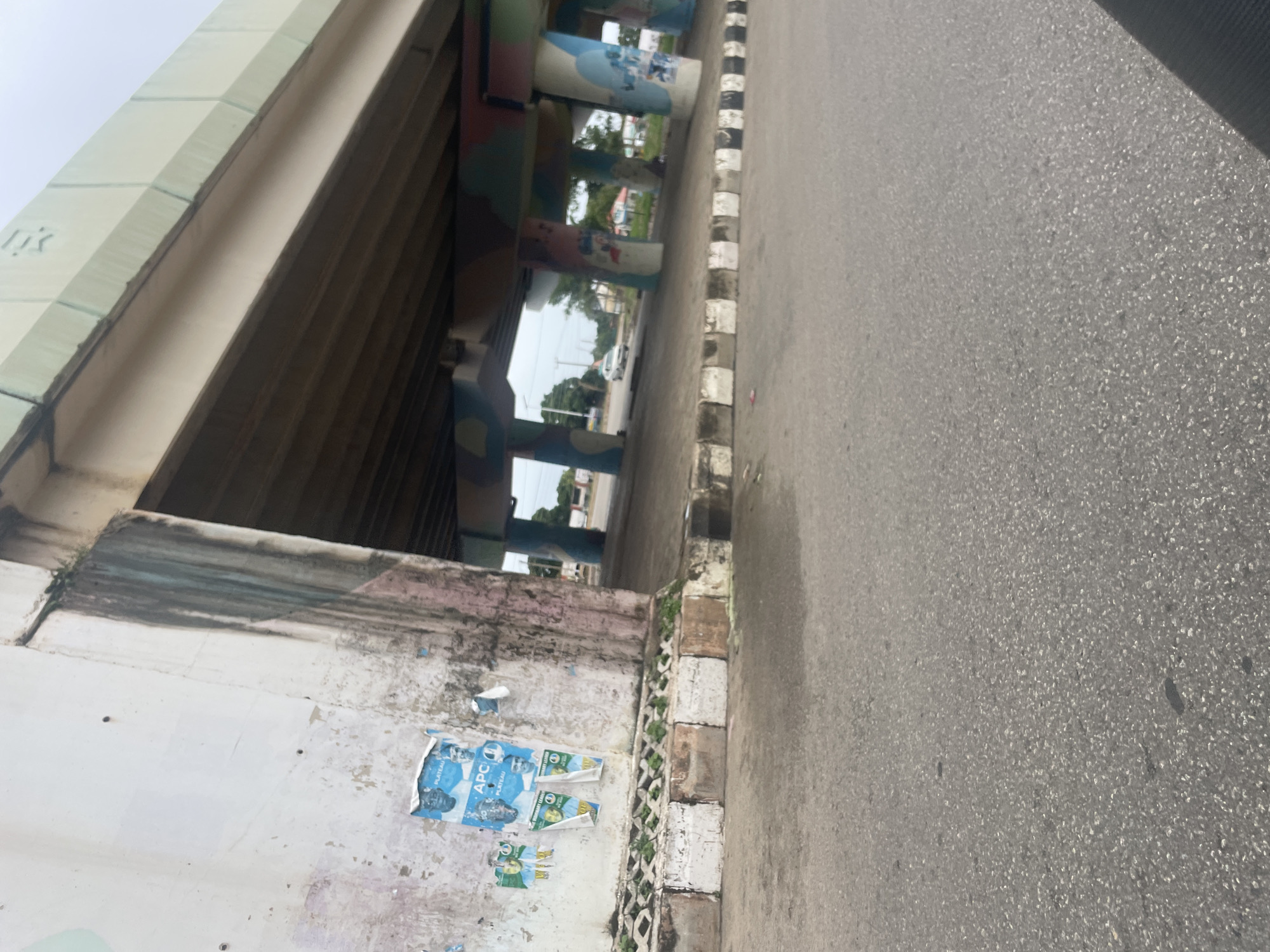 The image depicts an underpass beneath a concrete overpass in Jos, Nigeria. The concrete support pillars of the overpass are painted with colorful, abstract designs. On the left, a white concrete wall supporting the overpass has several torn political posters attached to it, some featuring images of men and the text "APC" and "PLATEAU." The ground beneath the overpass is paved and appears to be a road or parking area. A white car is visible in the background, driving under the overpass. The environment is outdoors with visible trees and utility poles in the distance, suggesting an urban or semi-urban setting. The pavement in the foreground is asphalt.