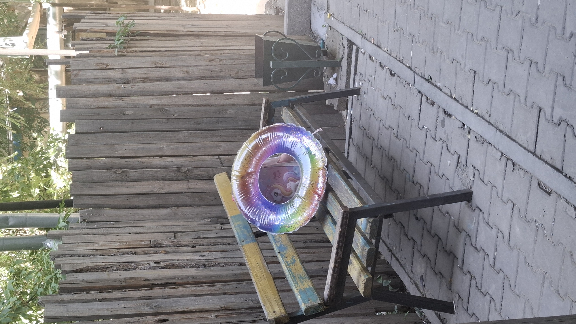 An outdoor scene captures a weathered wooden bench with a faded blue and yellow paint job, positioned in front of a rustic, vertical wooden plank fence. On the bench lies a circular, iridescent balloon filled with air, displaying a rainbow color gradient and a holographic glitter effect. Within the translucent balloon, an image of a unicorn is visible. To the right of the bench, a dark green, ornate metal trash receptacle stands on a paved area composed of interlocking grey concrete blocks. The background features green foliage and various pipes and structures, suggesting an urban or semi-urban setting. The lighting indicates it is daytime. No individuals are present in the image. The scene is likely located in Shymkent, Kazakhstan.