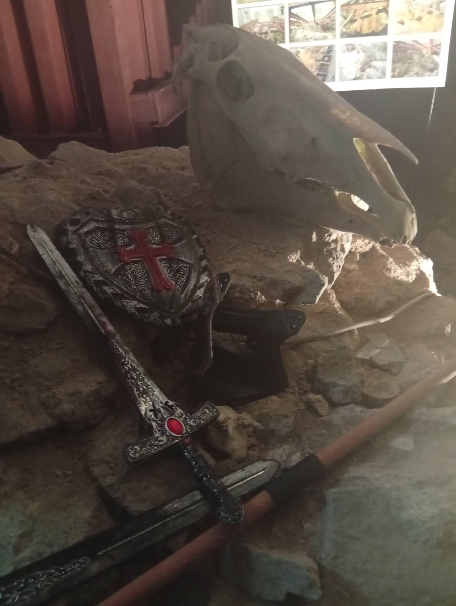 The image shows a still life arrangement of medieval-themed props, including a horse skull, a shield with a red cross, two swords, and an axe. The scene is set against a backdrop of rocks and sand. In the upper background, there is a display of multiple framed images, some appearing to depict historical or archaeological scenes. The lighting is dim, with a spotlight highlighting the central objects. The arrangement suggests a historical or fantasy exhibit, possibly within a museum or a themed attraction. The location is the Museum of the Earth in Sobótka, Poland, a place known for its geological and paleontological exhibits.