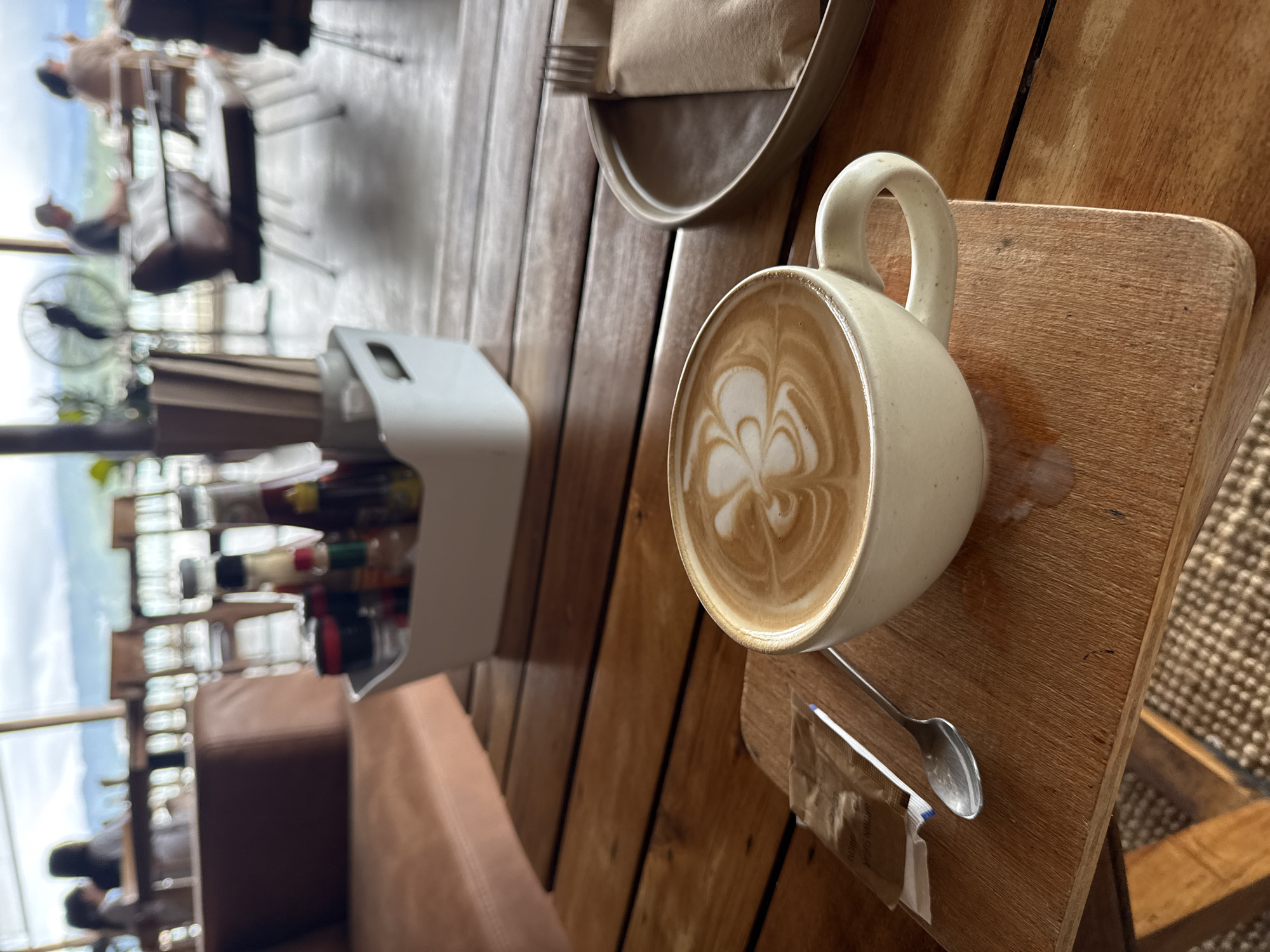A close-up shot captures a latte art coffee cup, a spoon, and a packet of brown sugar on a wooden table. The coffee cup, with its intricate latte art, sits on a small wooden board, which is placed on the table. A silver spoon rests next to the coffee cup, and a packet of brown sugar is positioned to the left of the spoon. In the background, out of focus, there are hints of a restaurant setting with people seated at tables, suggesting it might be a cafe or dining establishment. The overall ambiance suggests a relaxed moment, possibly at a location in Ko Samui District, Thailand, given the tropical-like lighting and open-air feel implied by the background elements.