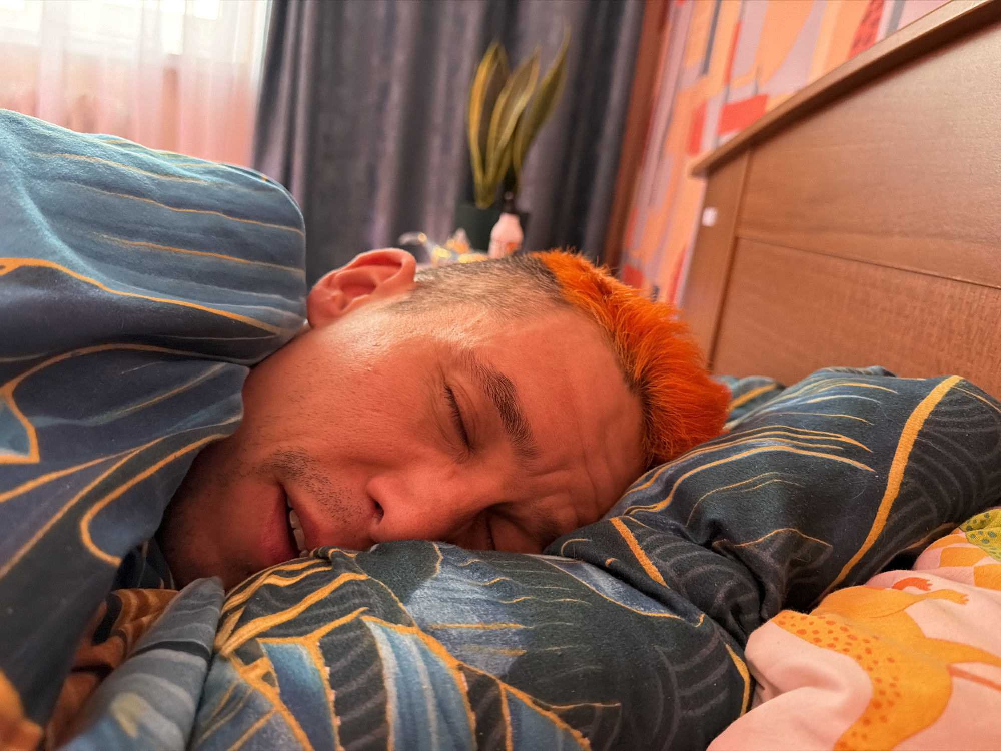 A man with orange hair is sleeping in a bed covered with patterned bedding. He is lying on his side with his eyes closed and a slight smile on his face. The bedsheets are predominantly blue and green with a yellow leaf pattern. Behind him, there is a wooden headboard, a curtain, and a plant on a table. The location is a bedroom in an apartment in Chaikovsky, Russia.