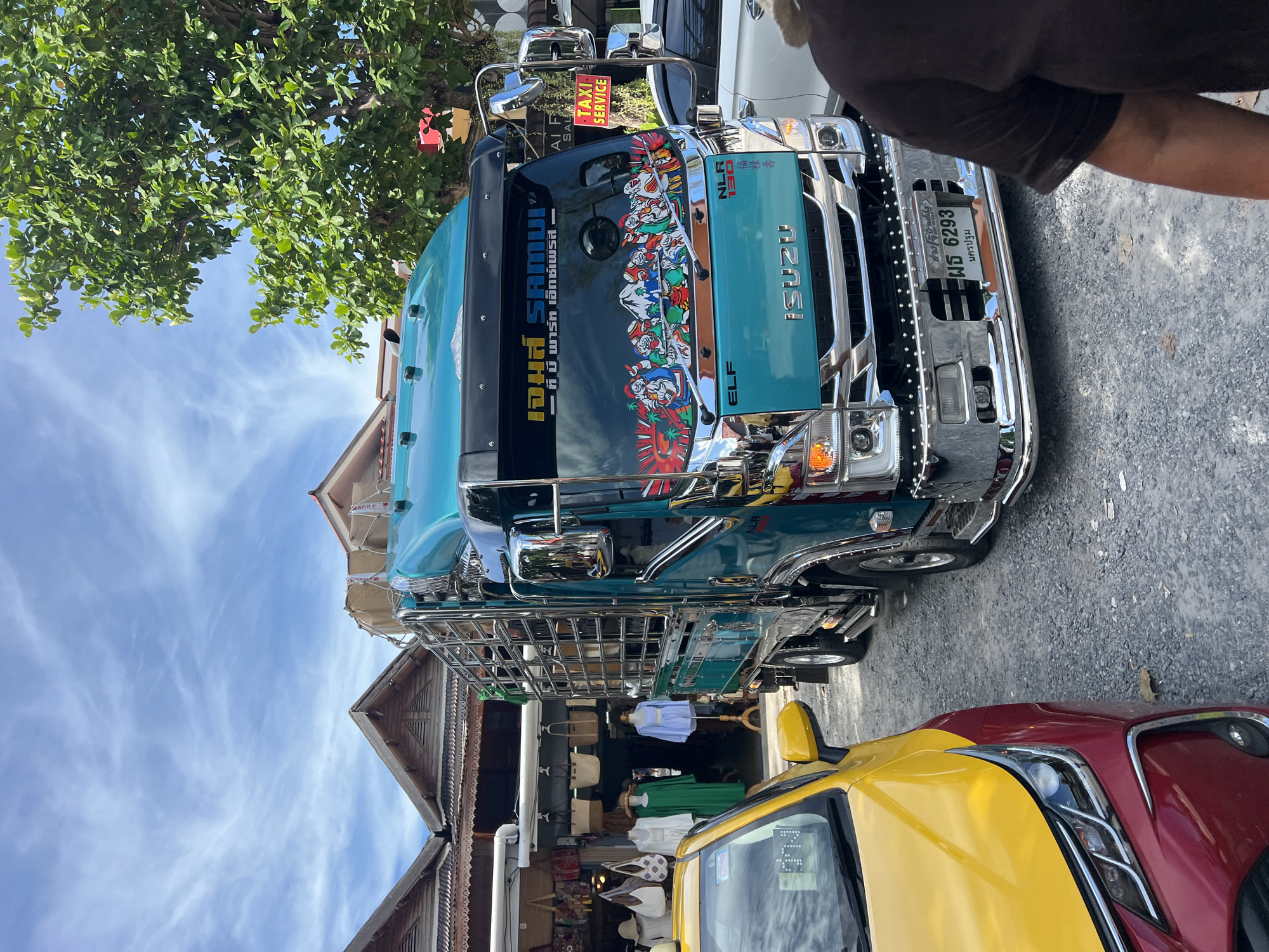 In Baan Plai Laem, Thailand, a teal Isuzu ELF truck is parked on a gravel road in front of a row of shops. The truck is decorated with chrome accents and colorful stickers on its windshield, which feature images of mountains and figures. A sign on the windshield reads "โจ้เ ร์ก Samui" and "บ้ าา วา ร ็ ค เฮ ็ ก เซ ็ ร ์ ว ิ ส ". A red and yellow taxi is parked to the left of the truck. The shops behind the truck display clothing and bags. A large green tree with dense foliage is visible to the right of the truck, and the sky is a mix of blue with wispy white clouds.