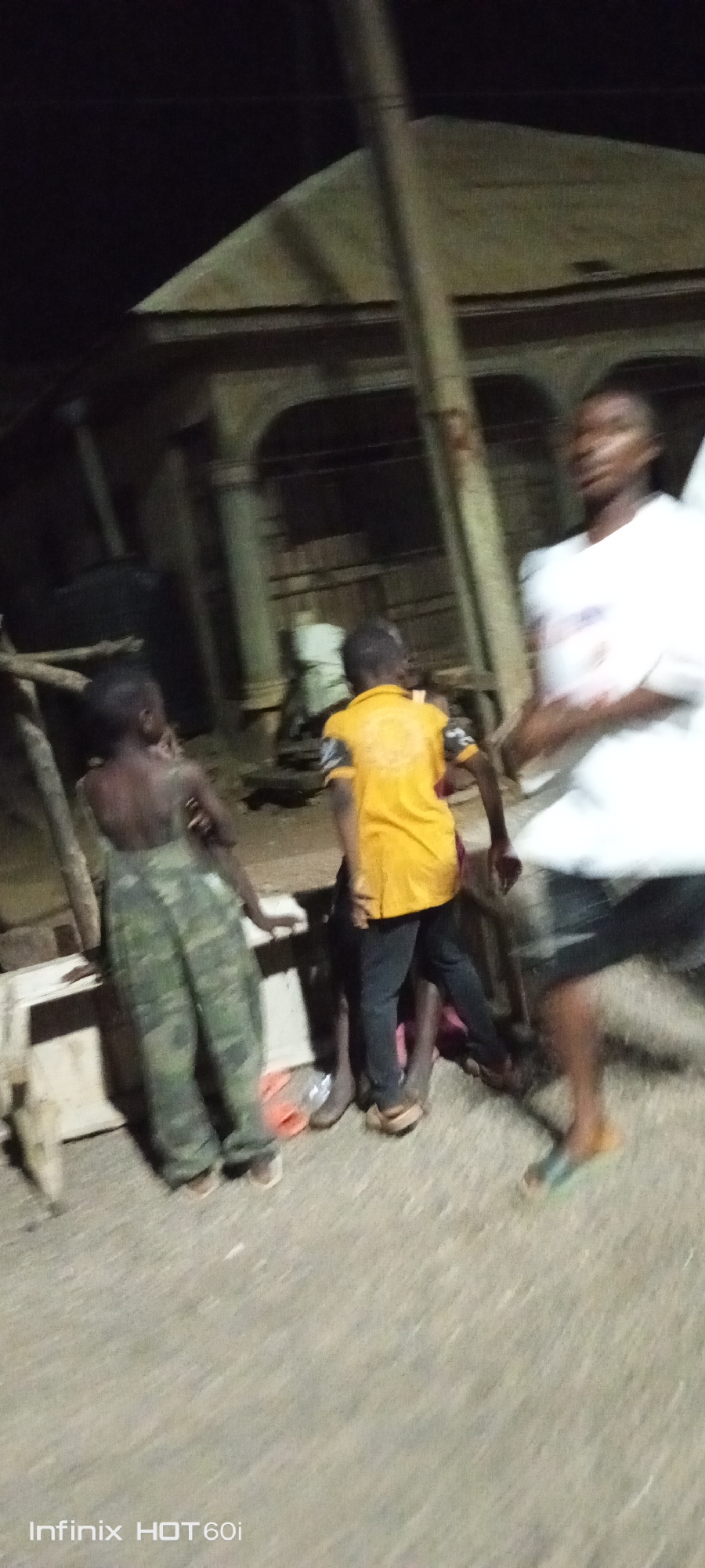 The image shows a group of individuals in Adankolo, Nigeria, at night. There are four or five individuals visible. One individual is wearing camouflage trousers and is shirtless. Another is wearing a yellow shirt and black trousers. Another individual is wearing a white t-shirt and black shorts. The individuals are standing in what appears to be a yard or open area in front of a building. The building behind them appears to be a structure with columns. The lighting is poor, with an Infinix HOT 60i watermark visible in the bottom left corner. There is some miscellaneous debris on the ground.