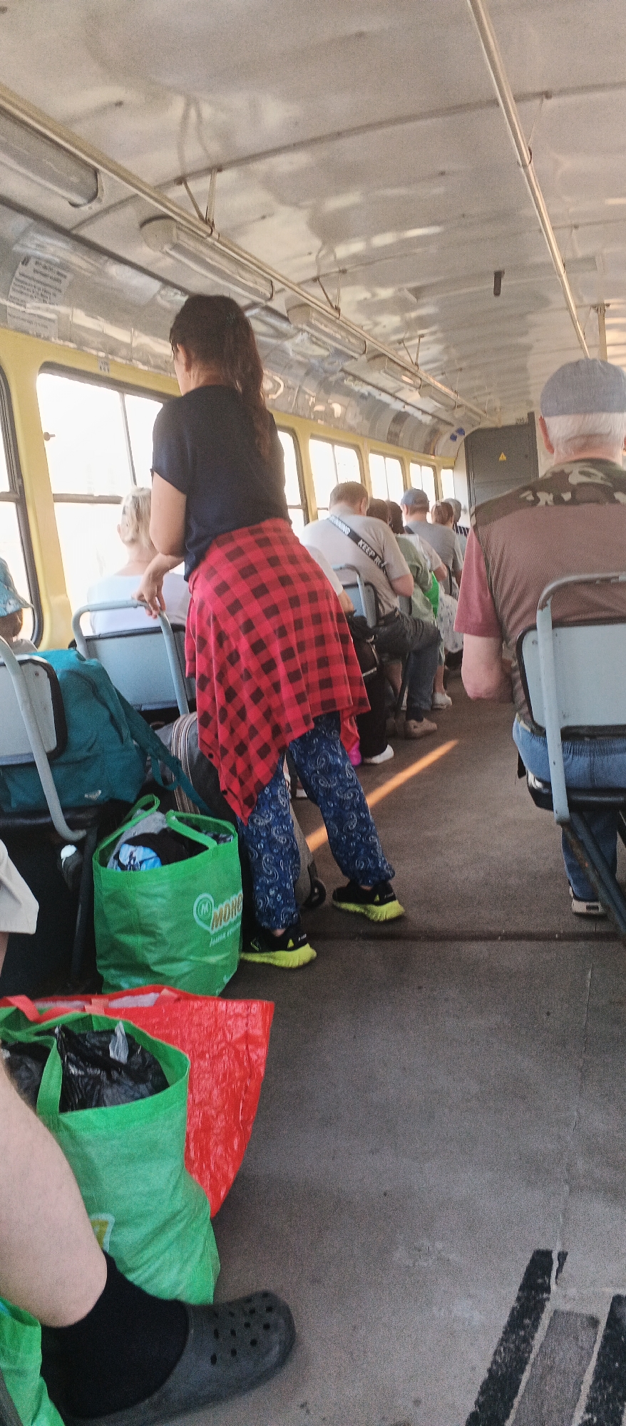 This is an eye-level shot taken inside a public transportation vehicle, possibly a trolleybus, in Izhevsk, Russia. The interior is utilitarian, with rows of blue and gray plastic seats. Sunlight streams in through large windows, illuminating the worn concrete floor. A woman with dark hair pulled back in a ponytail stands in the aisle, wearing a black t-shirt and blue patterned pants. A red and black checkered shirt is tied around her waist. Her stance suggests she is either standing or in the process of sitting down.
Several passengers are seated, facing forward, with their backs to the camera. One man wears a grey cap and a camouflage-patterned vest over a maroon t-shirt. Another man has a white t-shirt with black lettering that appears to read "KEEP R..." and a grey strap across his shoulder. Most passengers are dressed casually.
The floor is cluttered with various bags. A prominent bright green reusable shopping bag with a circular logo containing the word "MONO" is positioned near the woman's feet. It is filled with dark items, possibly clothing. A red plastic bag is also visible, along with another green bag containing a black trash bag. To the left of the frame, a person's leg wearing a black sock and a dark gray croc-style shoe is visible. The overall scene depicts a typical commute, with passengers and their belongings occupying the interior space.