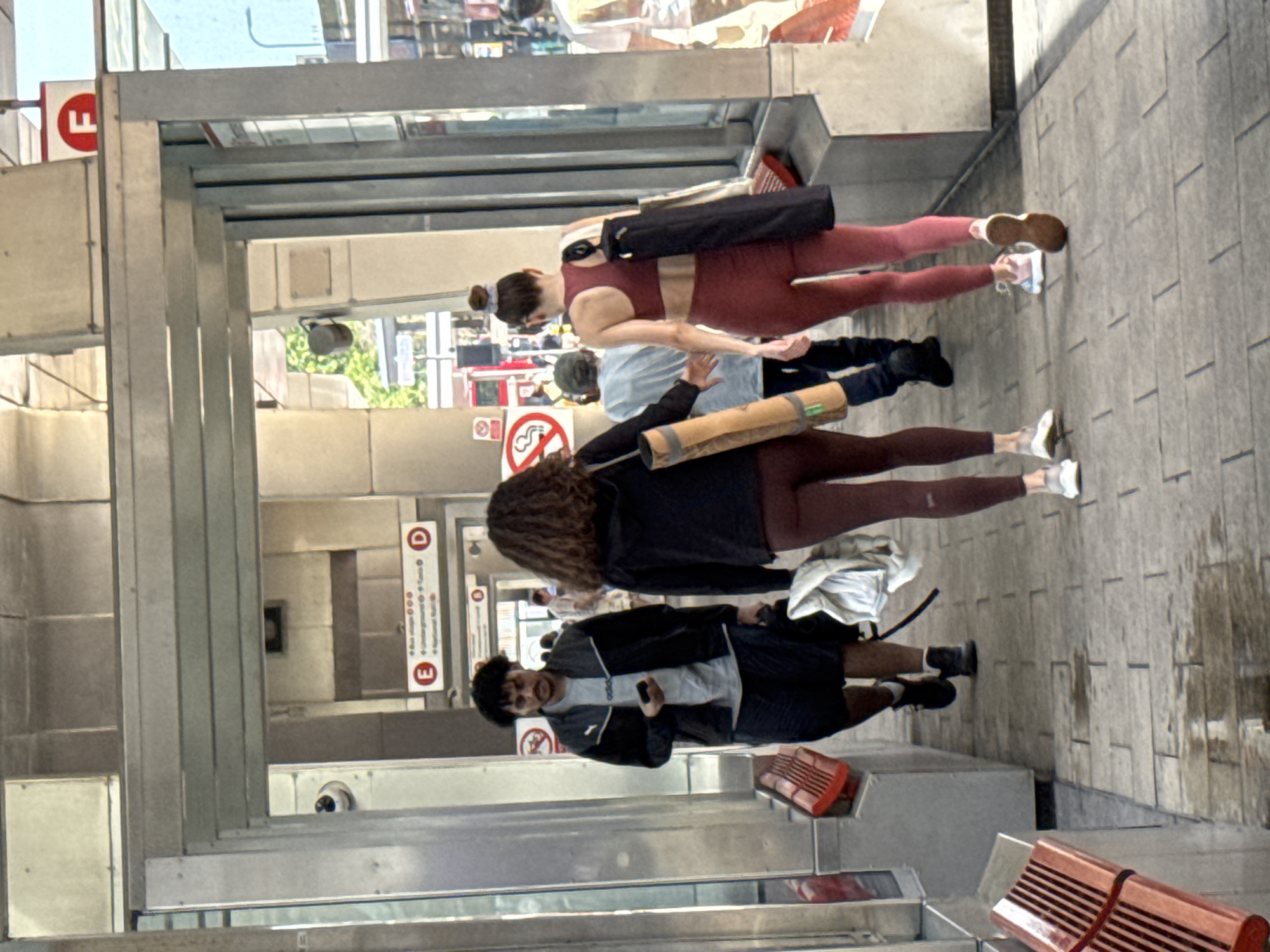 This image depicts a street scene in London, United Kingdom, possibly near a public transport station, as indicated by signage that includes the letter "E" and "D" along with some illegible text. Several individuals are present, walking or standing. In the foreground, a person is seen from the waist down, wearing a black jacket, grey t-shirt, and dark shorts. They are holding a black object, possibly a phone. To their left, a red trash can is visible. Further back, a woman in maroon athletic wear with a black yoga mat slung over her shoulder walks away from the camera. Another person in a light blue shirt is also walking away. A person in a dark jacket and maroon leggings is holding a rolled-up mat. The ground is paved with large tiles. The lighting suggests it is daytime, and the weather appears overcast, with wet patches on the ground indicating recent rain. The overall atmosphere is that of a busy urban environment.