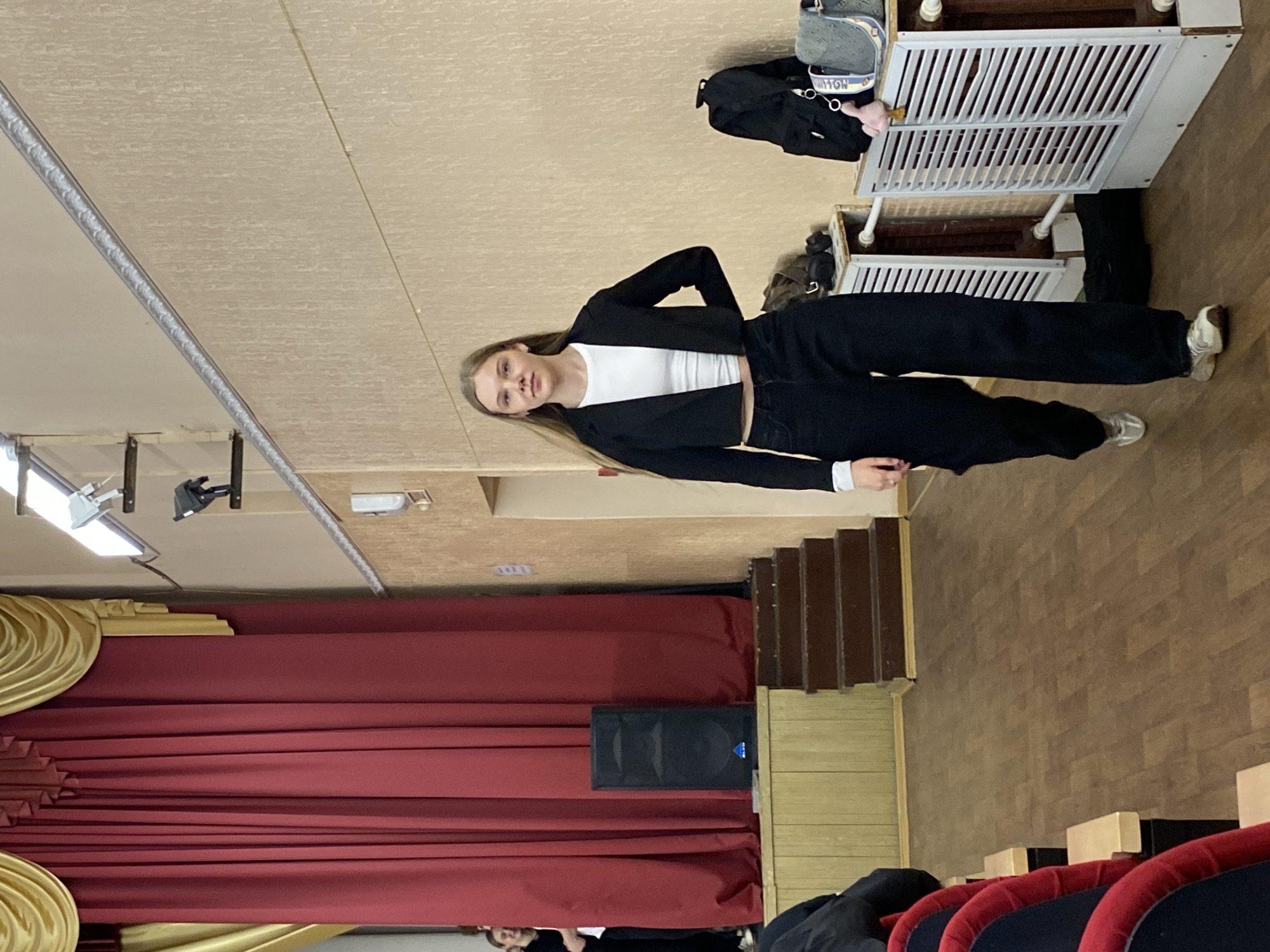 The image, originally oriented 90 degrees counter-clockwise, displays a young woman standing indoors, likely in an auditorium or assembly hall.
The central subject is a young woman with long, straight light brown hair, dressed in a black cropped blazer over a white crop top, black wide-leg trousers, and white sneakers. She stands with a neutral expression, looking directly at the camera.
In the background to her right, two white, slatted furniture pieces, resembling cribs or playpens, are stacked. On the upper one, a gray-blue patterned bag with the partial text "VUITTON" is visible, along with a black item. The floor throughout much of the scene is light brown parquet or wooden flooring.
To the woman's left and extending into the background, large red stage curtains with golden, ornate valances frame a stage area. A black speaker, featuring a subtle blue logo, is positioned in front of these curtains. Brown wooden stairs are visible in the mid-ground, leading upwards. In the lower right foreground, the backs of red upholstered theater-style seats with dark armrests are partially visible, indicating an audience seating area.
The ceiling features modern light fixtures, including rectangular panels and a spotlight. The indoor setting, with artificial lighting, provides no specific cues for the time of day or weather. The woman appears to be posing for the photograph, with no other discernible activity taking place. The overall environment suggests a public or institutional venue in Votkinsk, Russia.