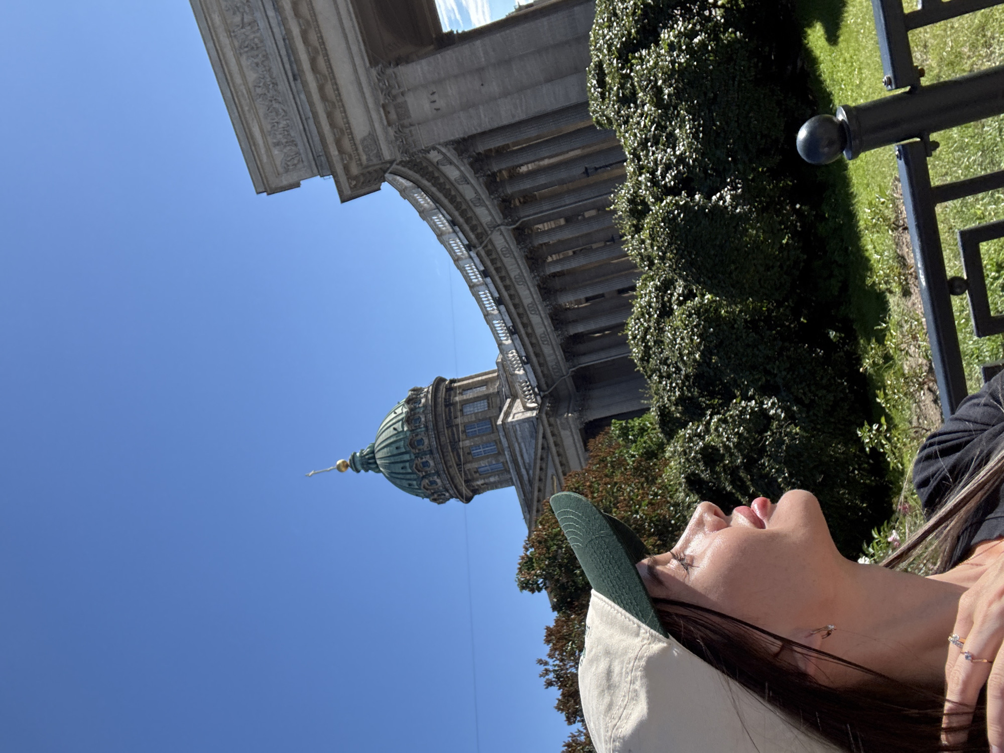 The image shows a woman wearing a beige and green baseball cap, looking up and to the left. Behind her, a grand architectural structure with a domed roof and colonnades is visible under a clear blue sky. The building appears to be Kazan Cathedral, a prominent landmark in Saint Petersburg, Russia. Lush green bushes and manicured lawns are in the foreground. The overall scene suggests a sunny day in an urban setting.